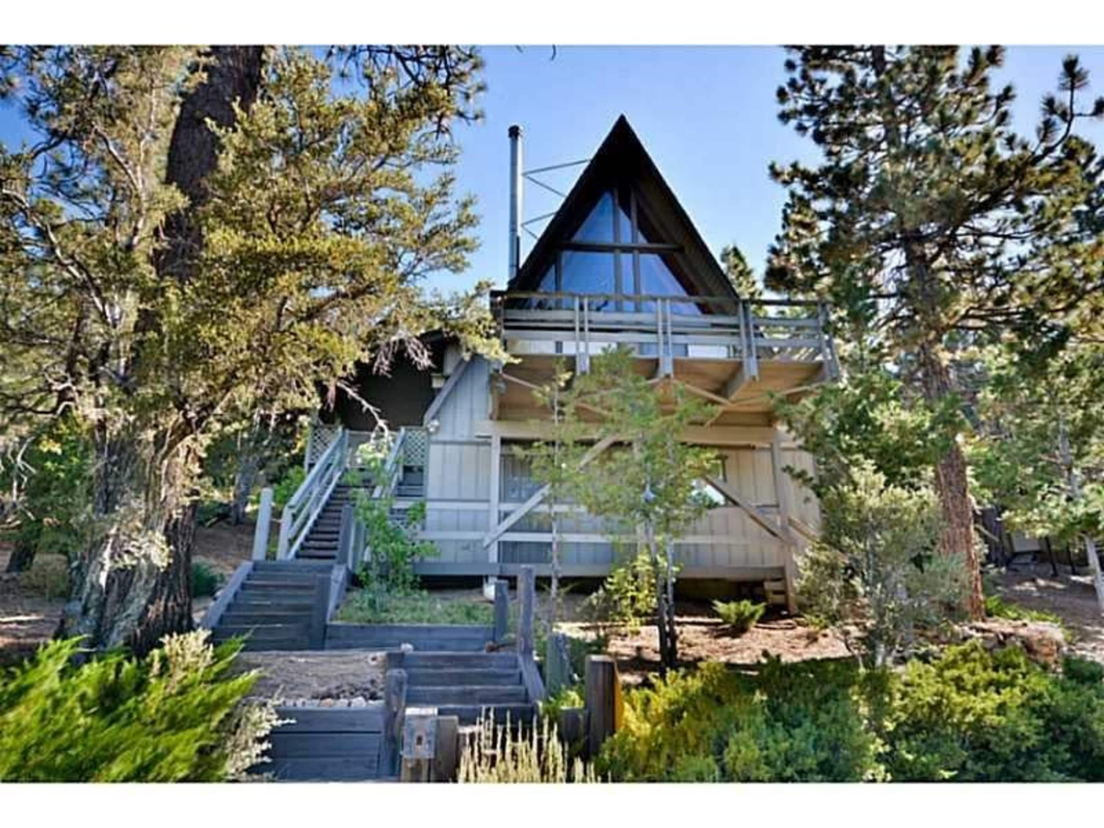 The image shows a two-story A-frame cabin nestled among tall pine trees, featuring a balcony and a wooden staircase leading to the entrance.