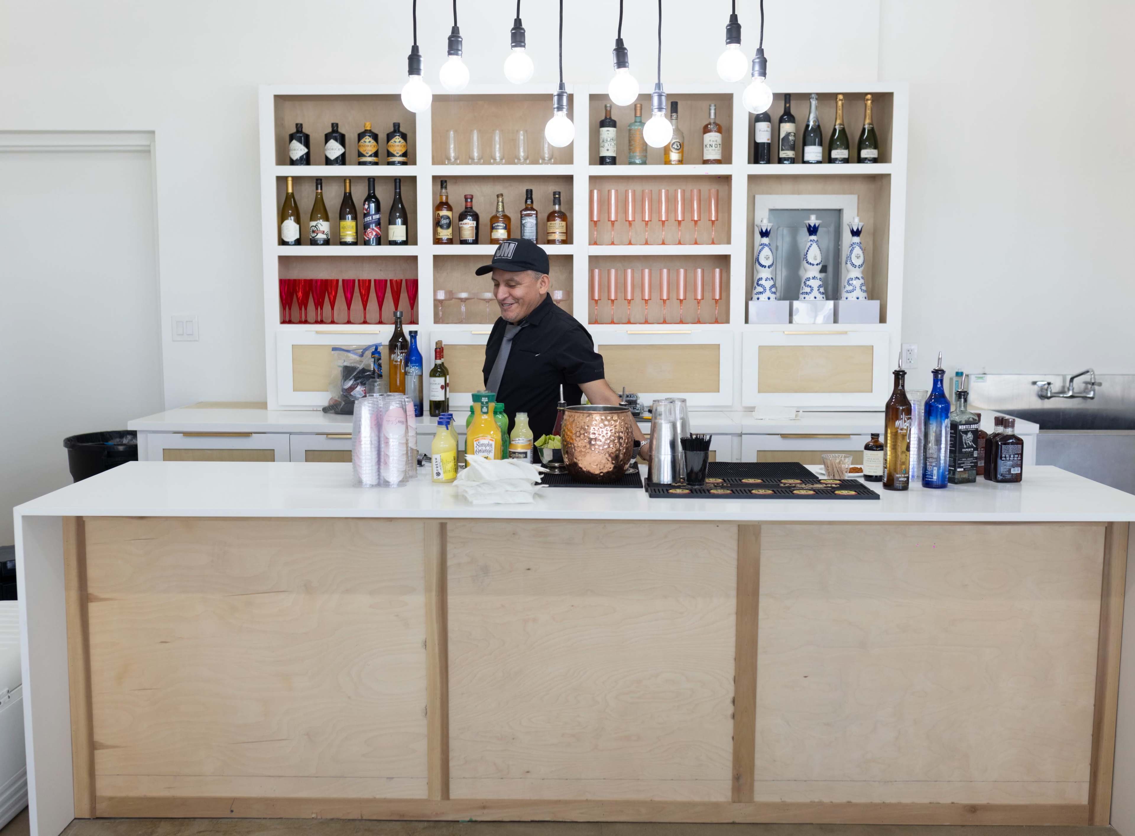 A bartender stands behind a sleek wooden bar filled with various bottles and drink supplies, with pendant lights hanging above.