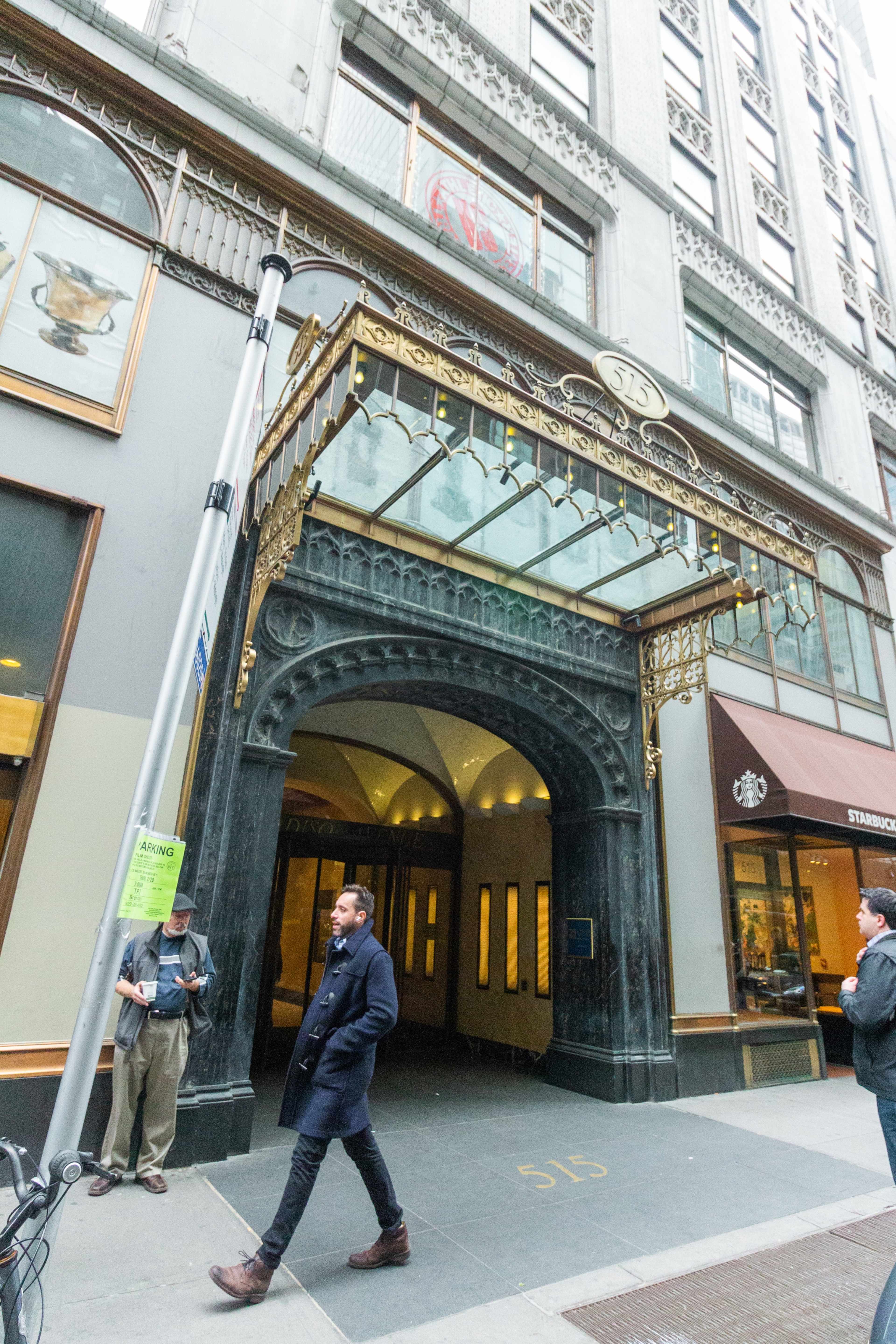 A person walks past a historical building entrance adorned with ornate architecture and a glass canopy.