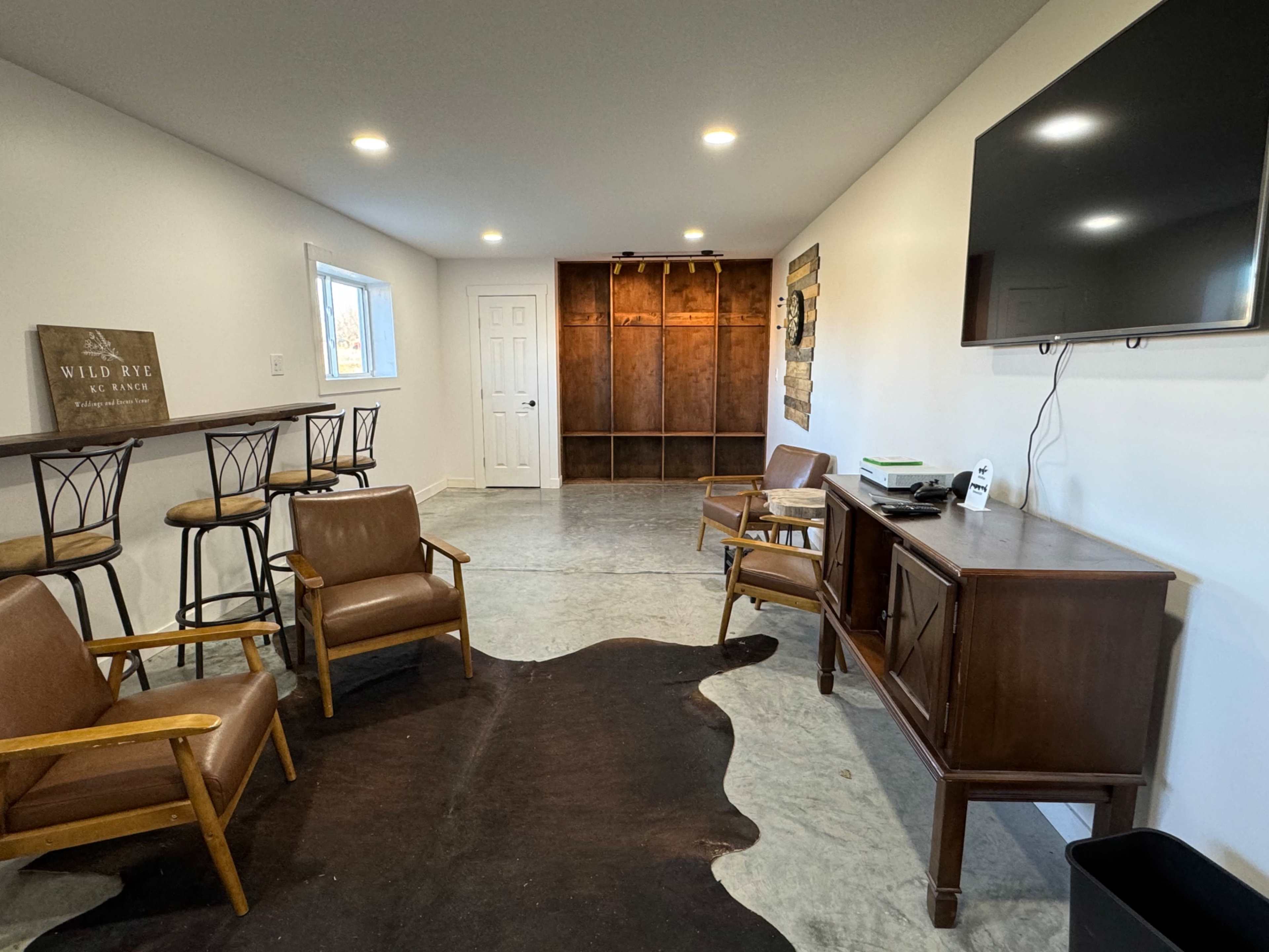 The image shows a spacious room with two bar stools, several chairs, a television mounted on the wall, and a wooden cabinet against one side, complemented by a large rug on the floor.