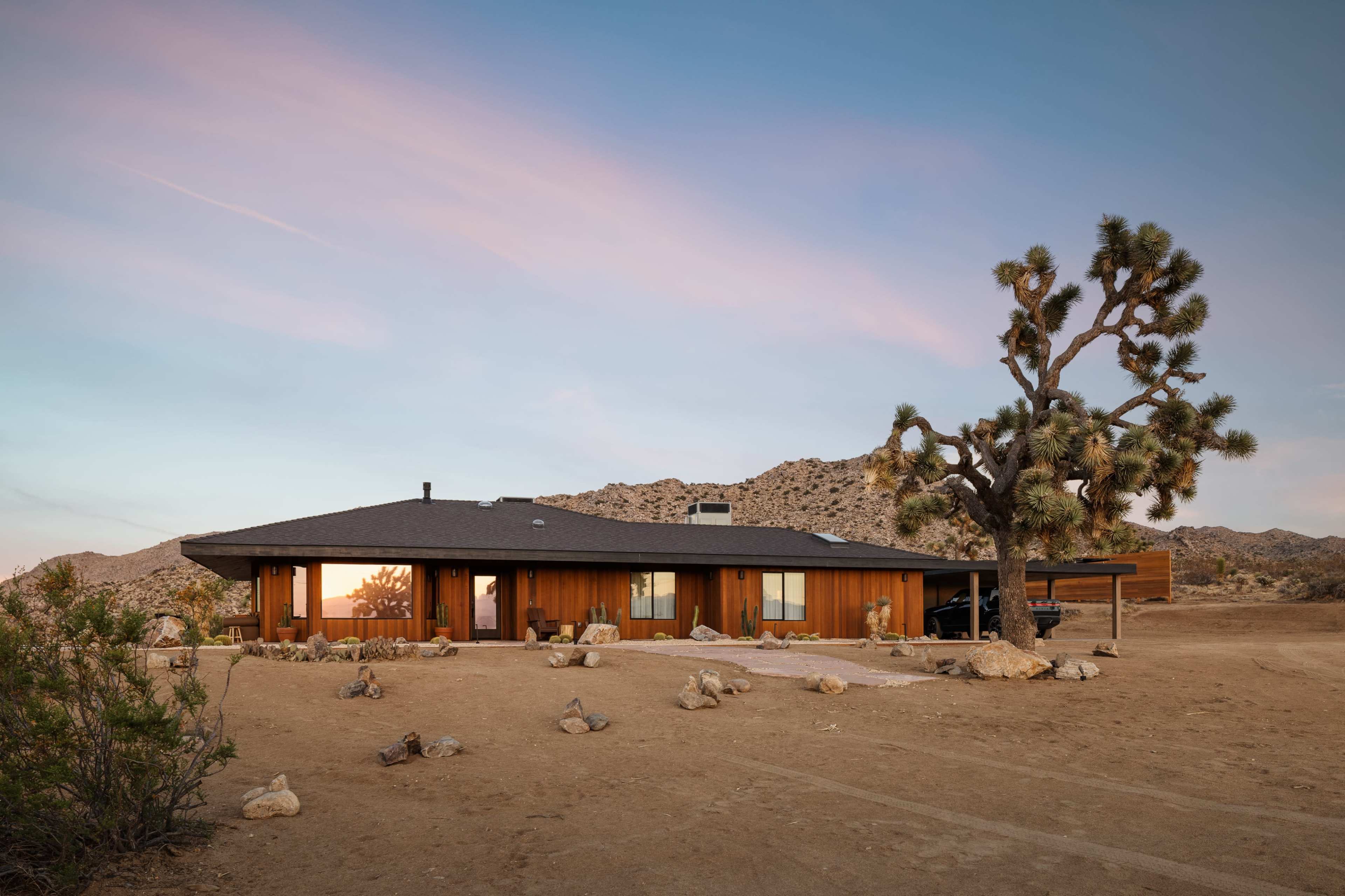 A mid-century modern house with wooden siding is situated in a desert landscape featuring large boulders and a Joshua tree in the foreground.