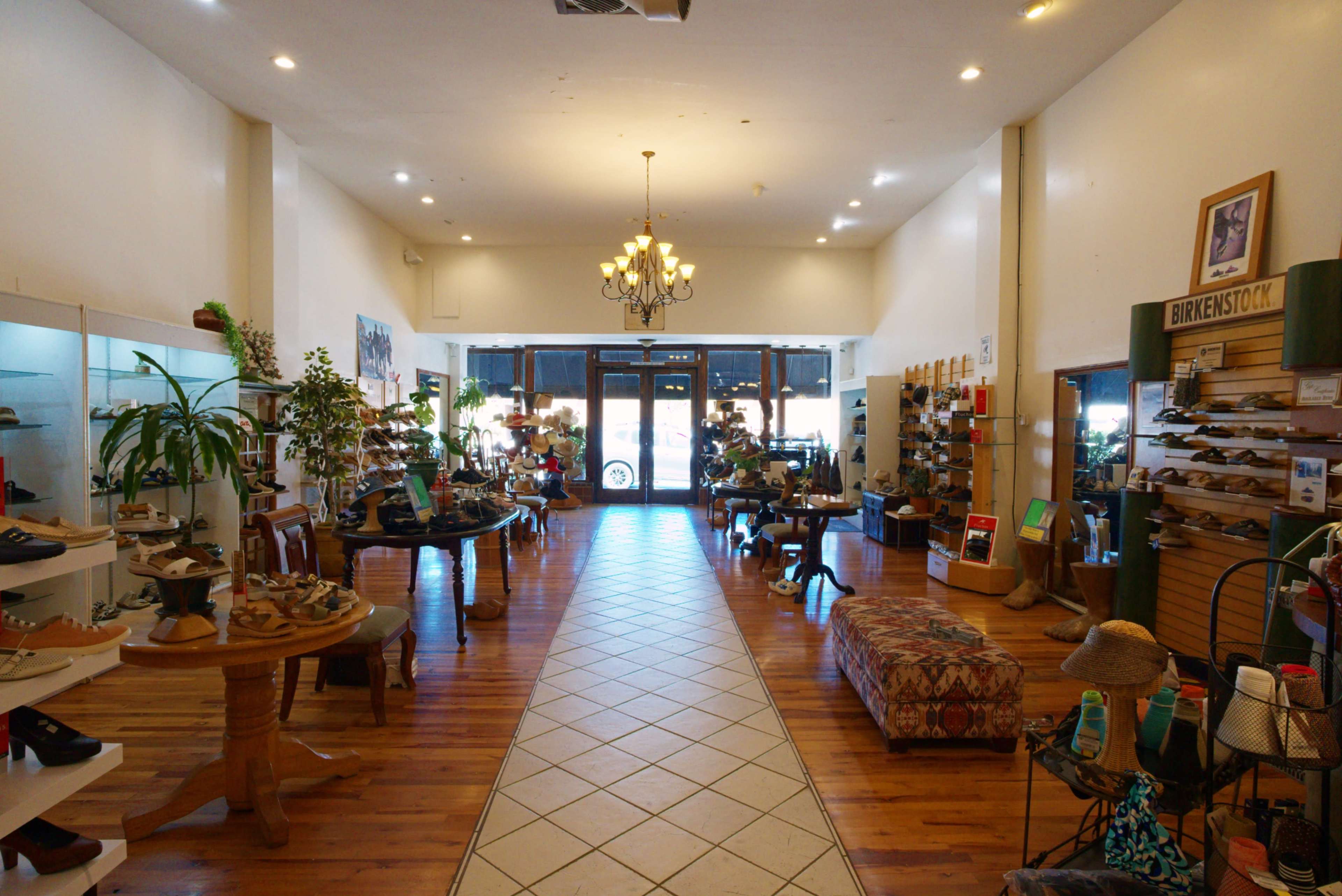 The image shows the interior of a shoe store featuring wooden flooring, displayed footwear on shelves, and a central seating area.
