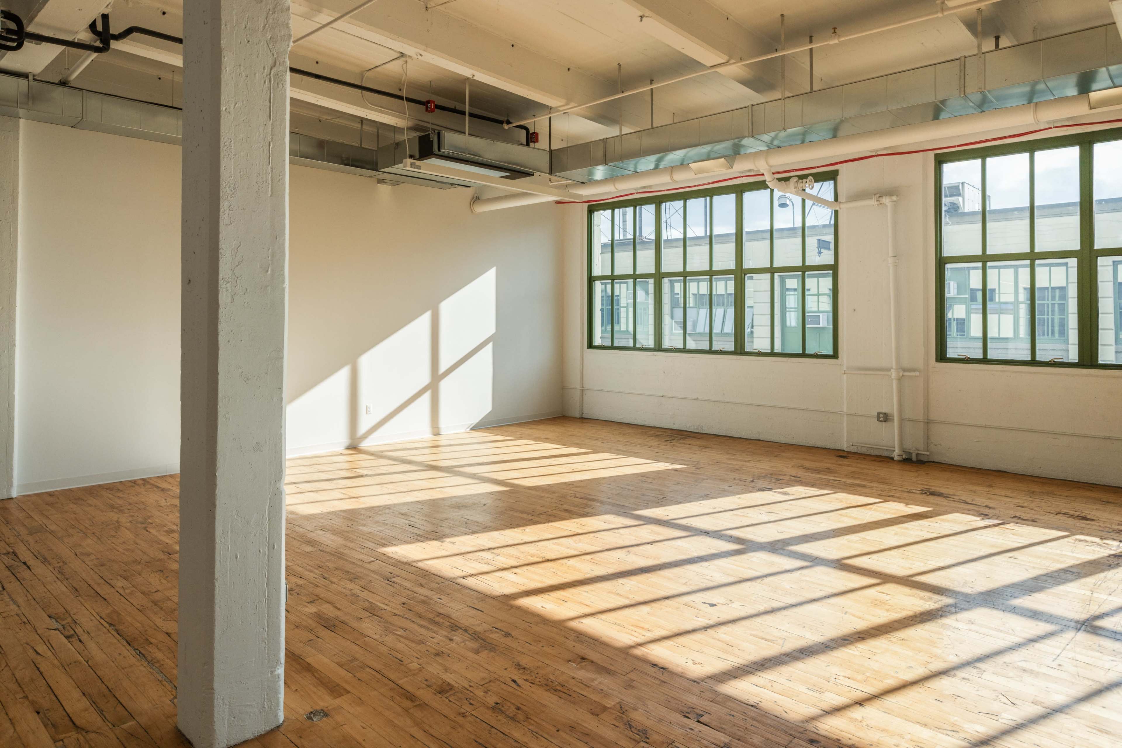 The image shows a spacious, vacant room with large windows allowing sunlight to cast shadows on the wooden floor.