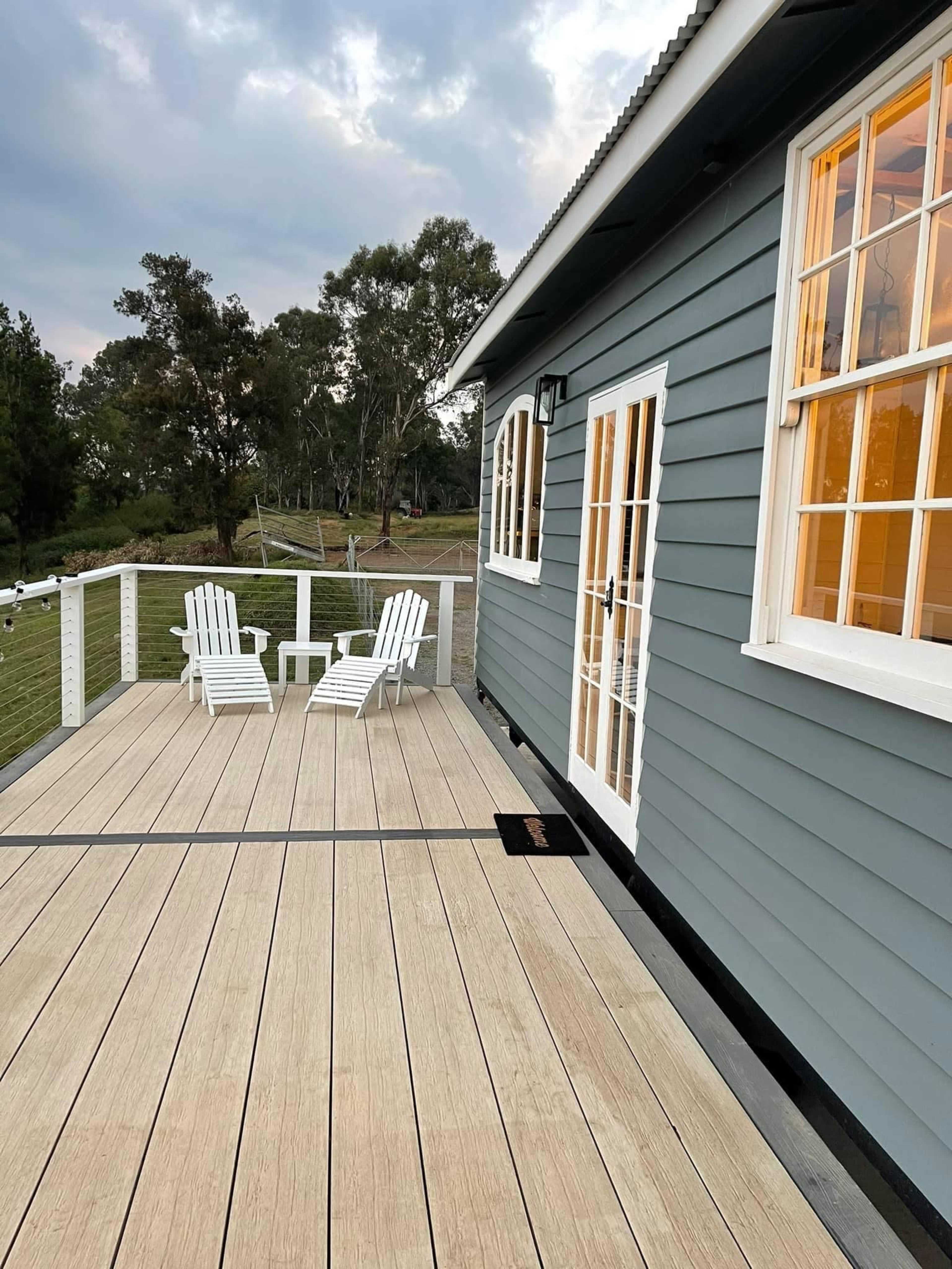 A wooden deck with two white chairs in front of a blue-gray house, and trees are visible in the background.