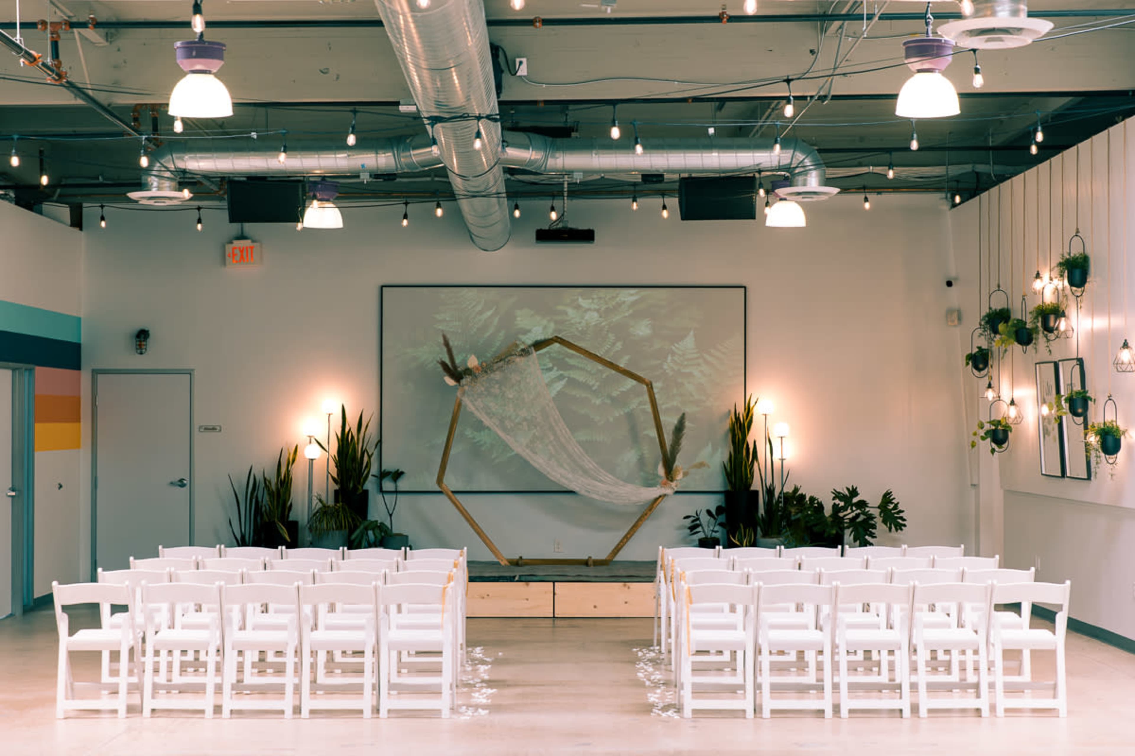 An indoor wedding setup with white chairs arranged in rows facing a decorated altar featuring a large geometric frame, backlit by soft lighting.