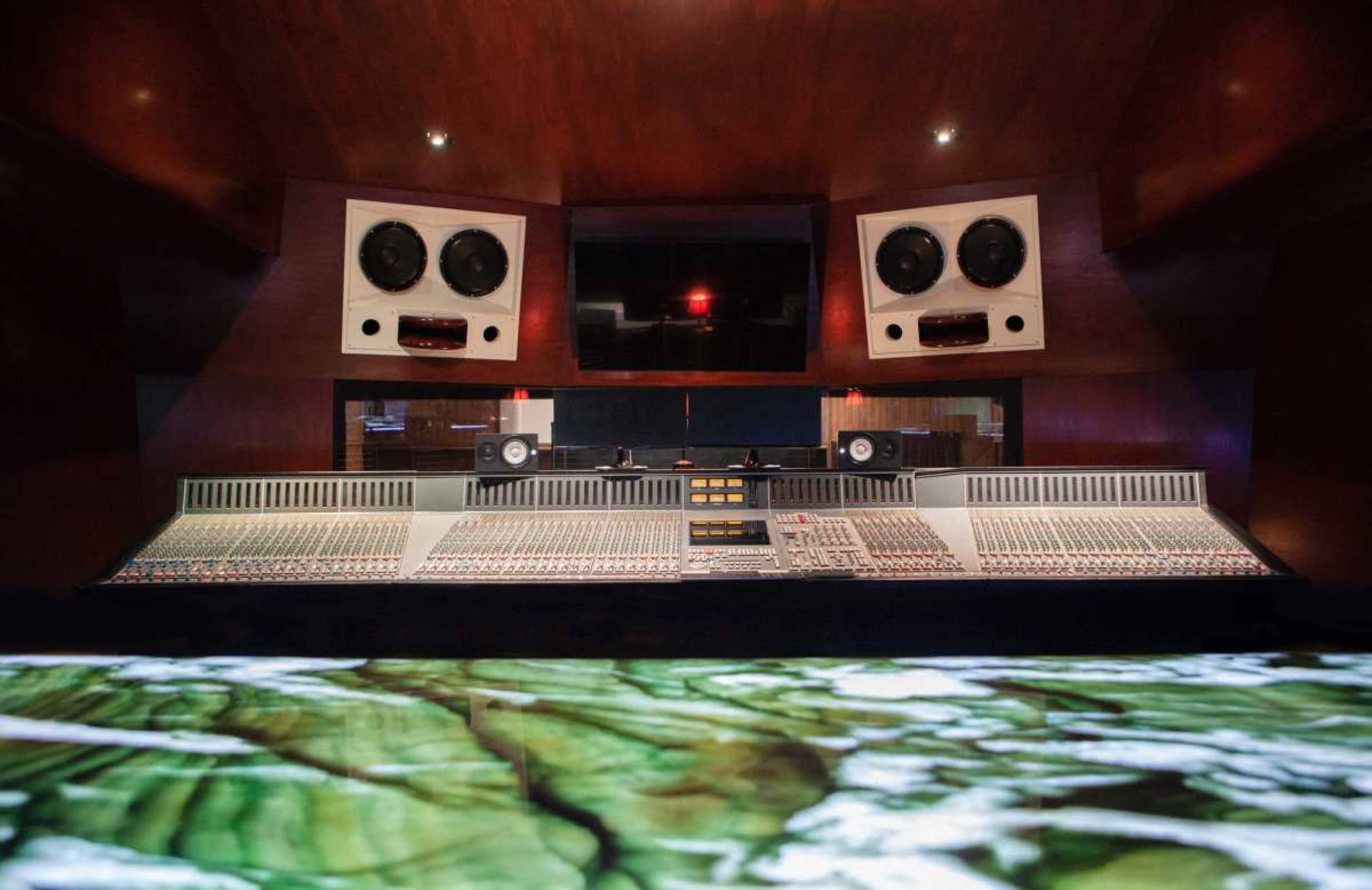 The image shows a modern music recording studio with a large mixing console, speakers, and a screen in a wood-paneled environment.