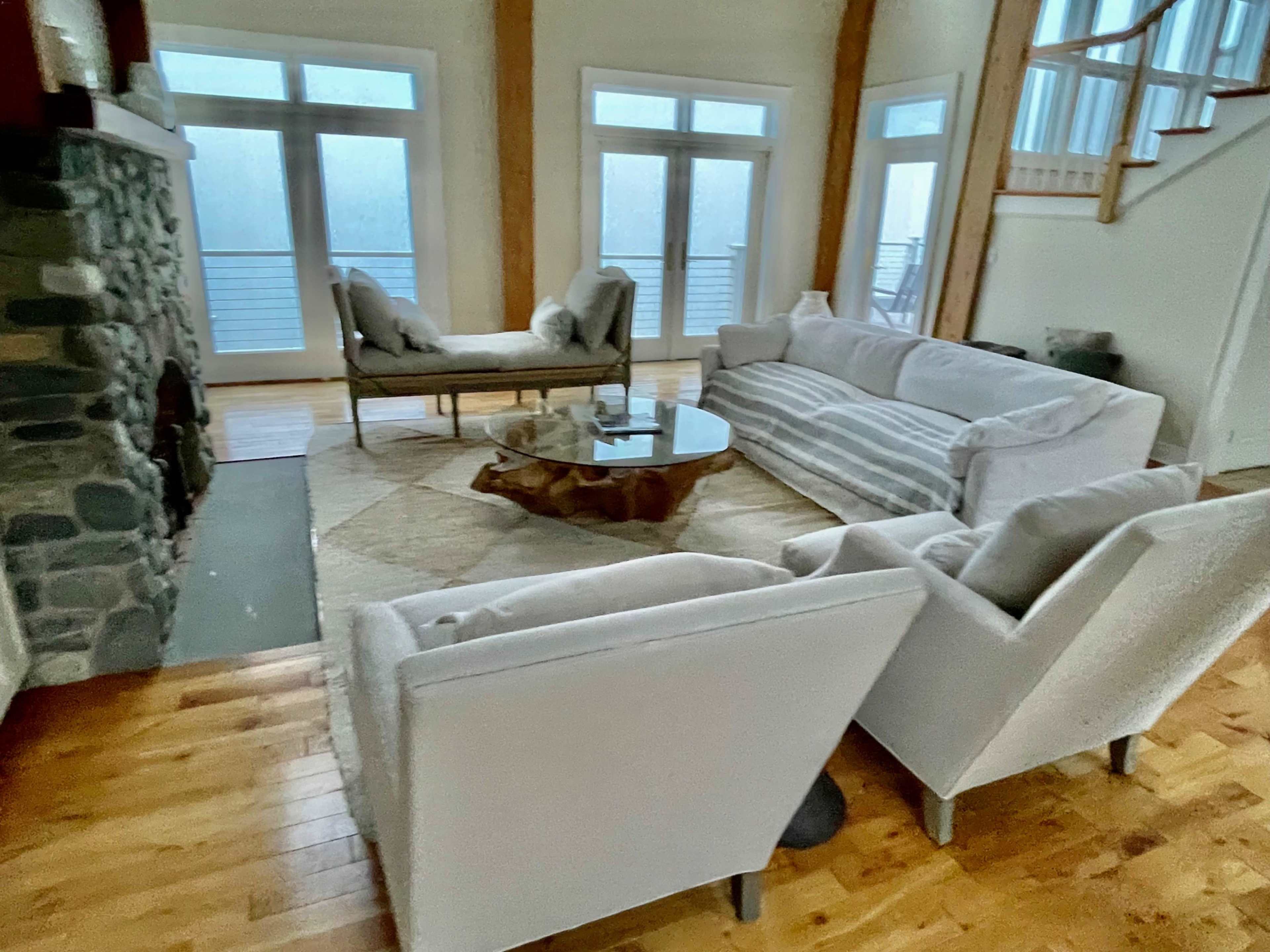 The living room features a stone fireplace, two armchairs, a striped sofa, and a round coffee table on a large area rug, with large windows providing natural light.