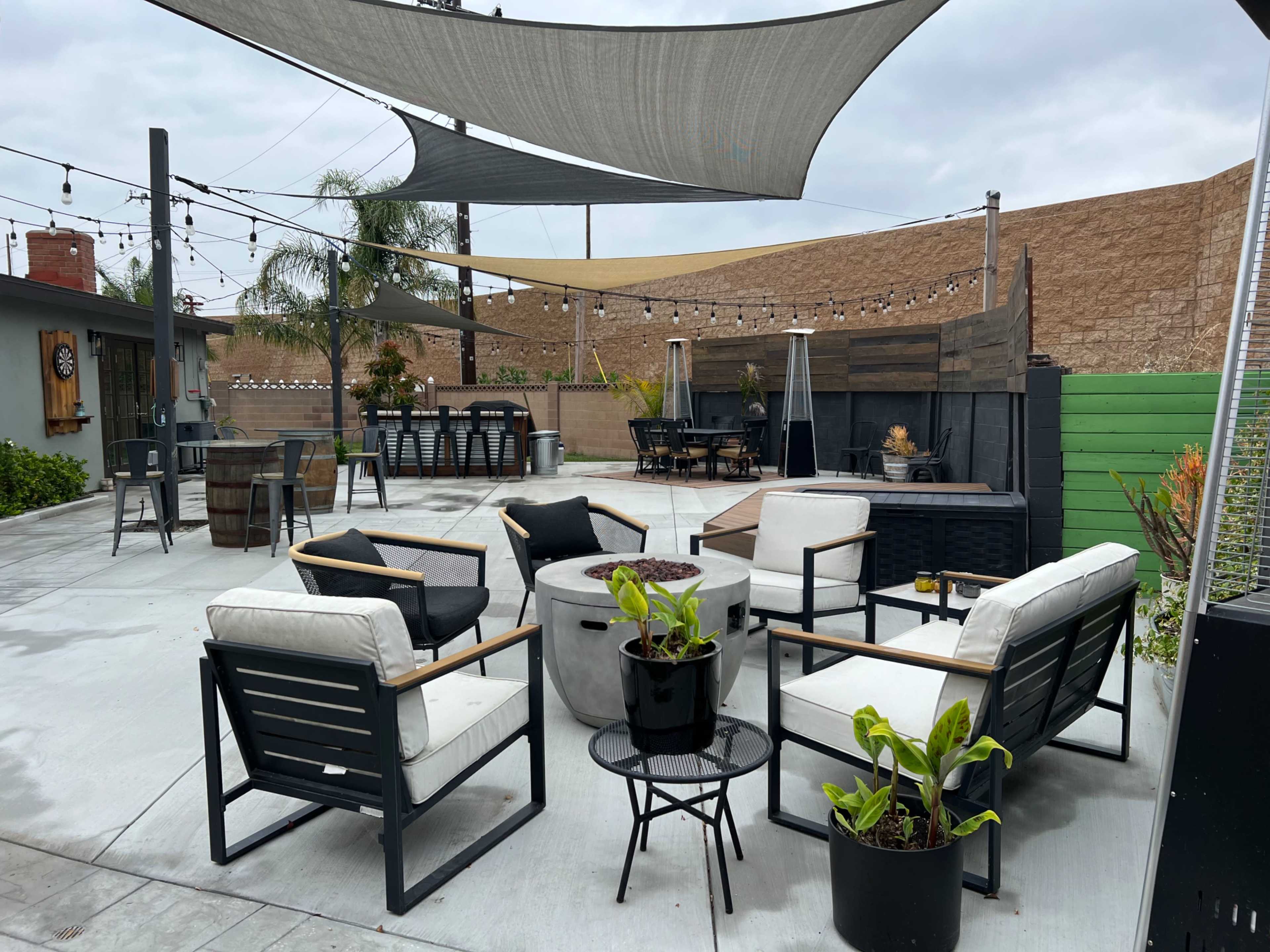 An outdoor patio area with modern seating, a fire pit, and shaded sections, surrounded by greenery and wooden fencing.