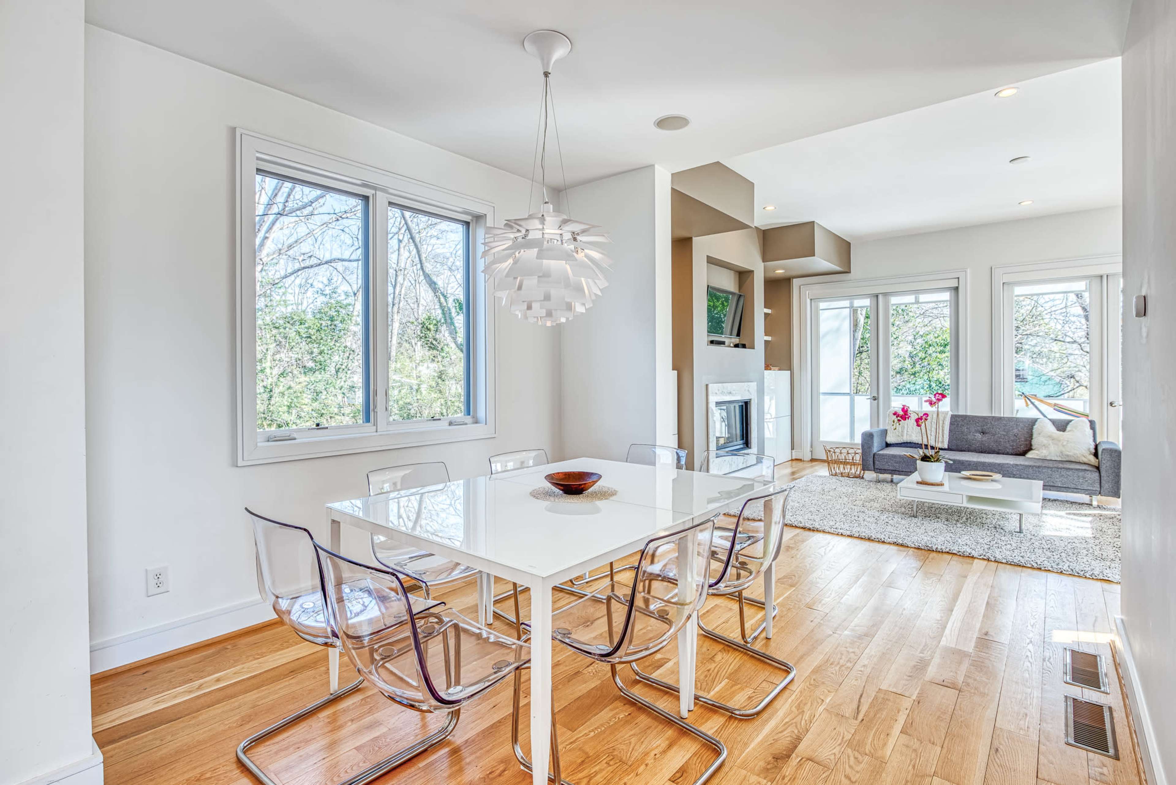 A modern dining area with a white table and clear acrylic chairs, illuminated by a distinctive pendant light, adjacent to a living space with large windows and a fireplace.