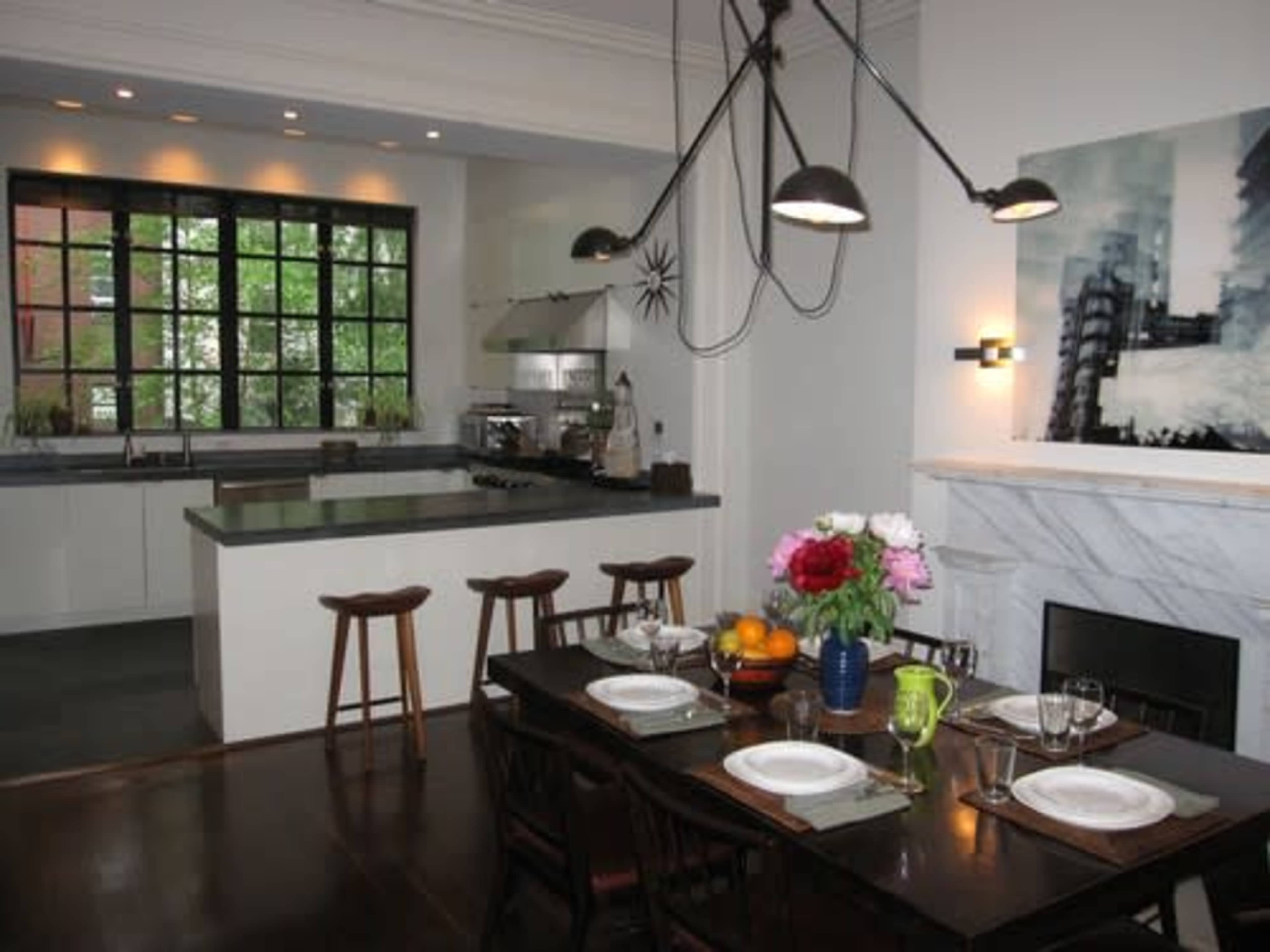 A modern kitchen-dining area with a rectangular dining table set for a meal, surrounded by chairs, and large windows allowing natural light to enter.
