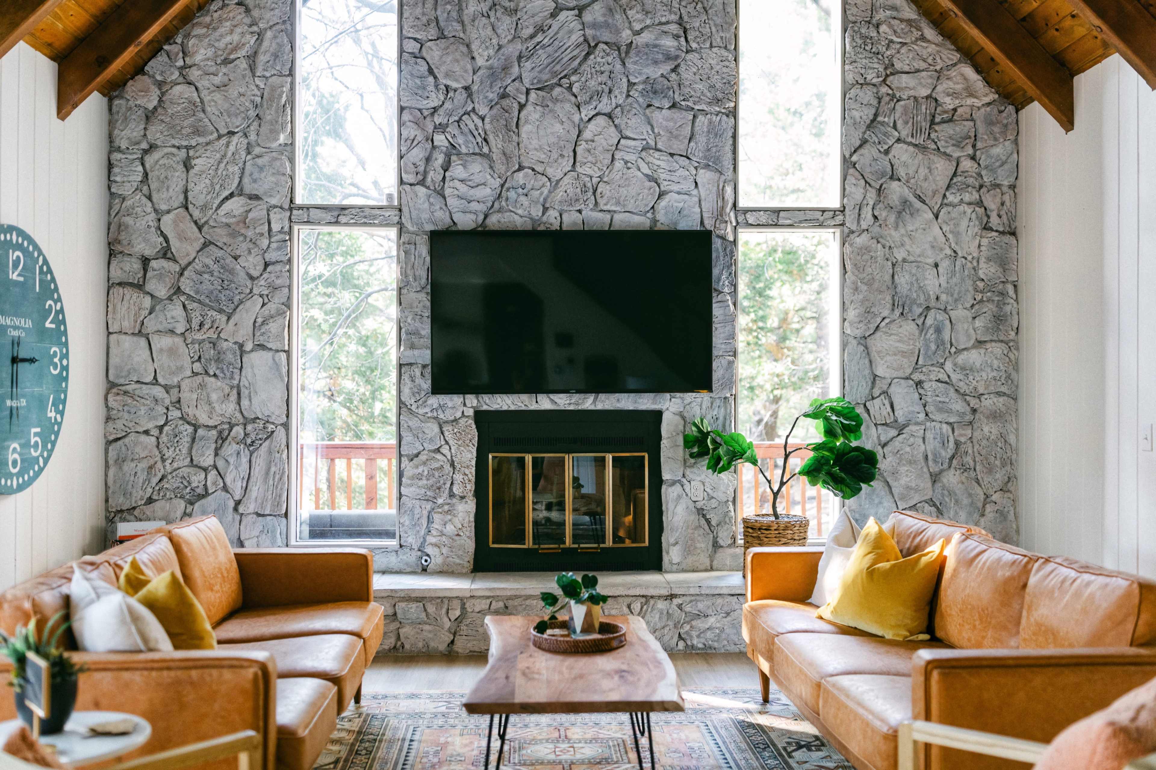 The living room features a stone fireplace with a mounted television above it, framed by large windows and furnished with two leather sofas and a wooden coffee table.