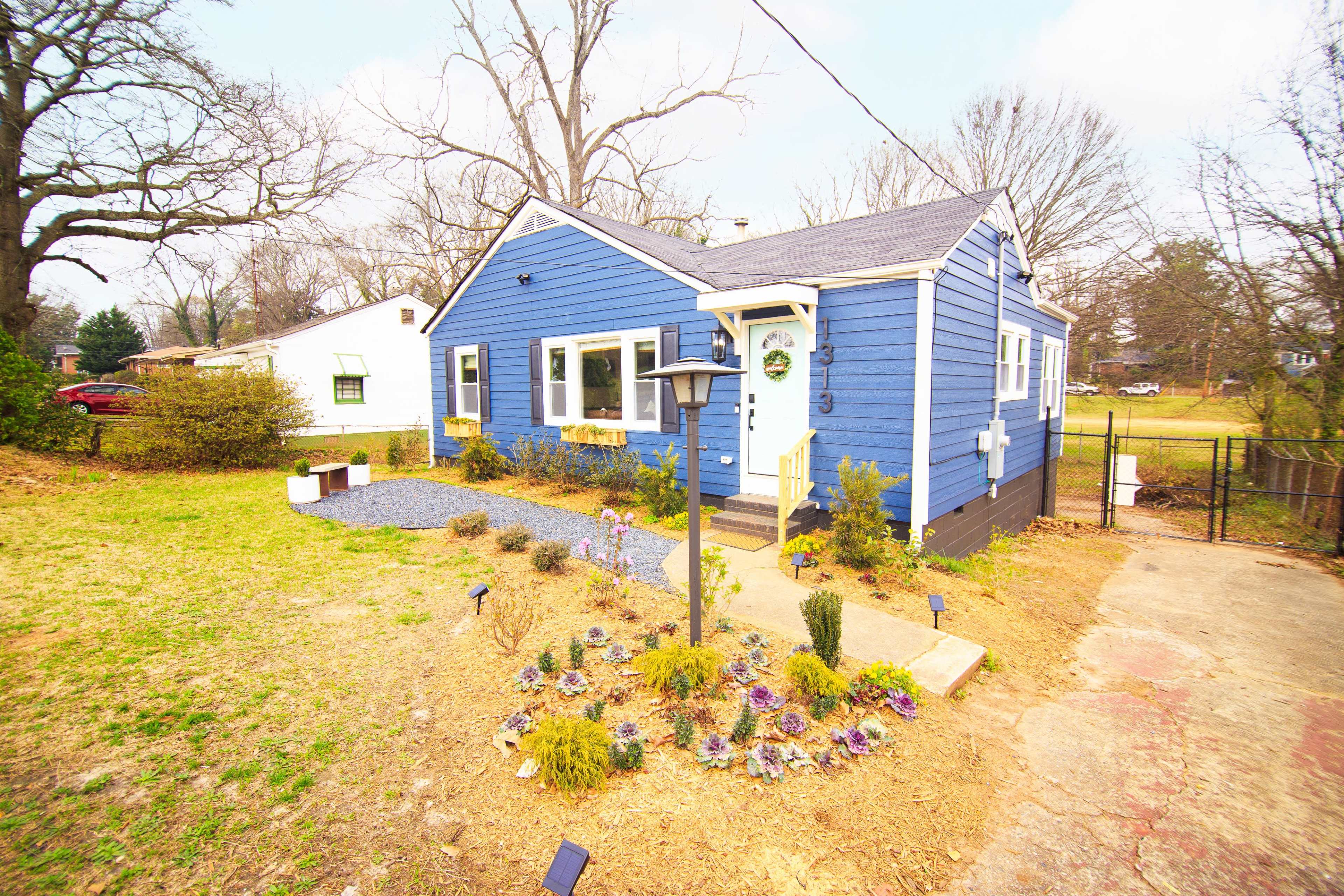 The image shows a blue clapboard house with a landscaped front yard featuring a pathway, flower beds, and a lamppost.