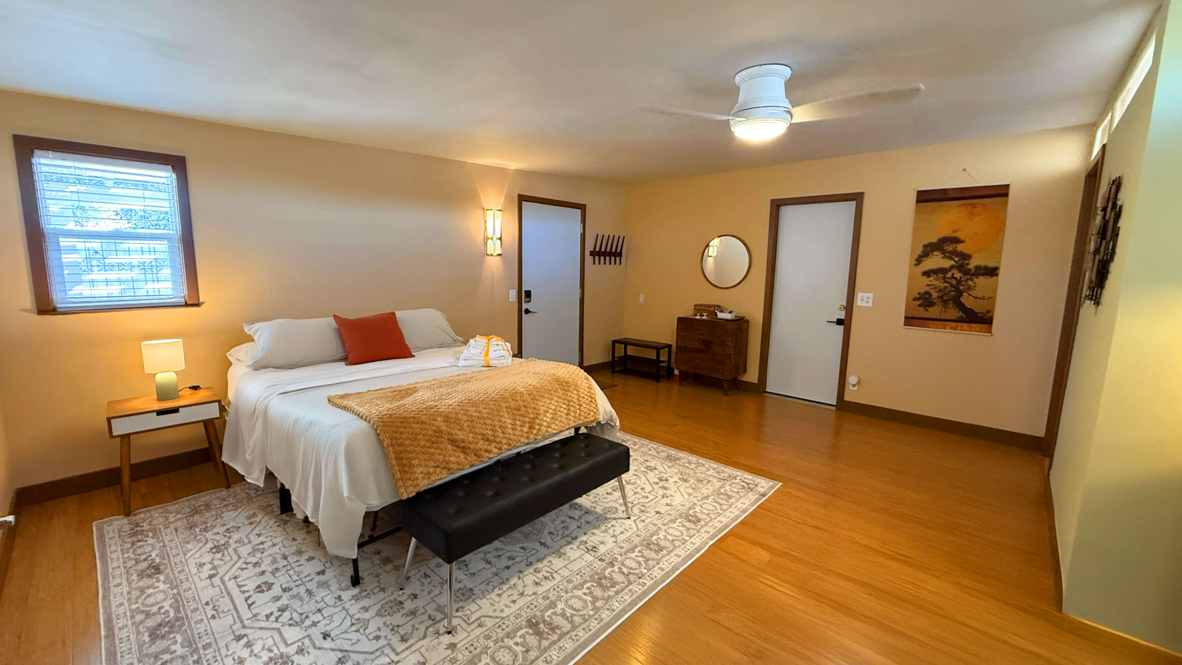 The image shows a spacious bedroom featuring a bed with white linens, a brown throw blanket, a nightstand with a lamp, and a vintage dresser against the wall.
