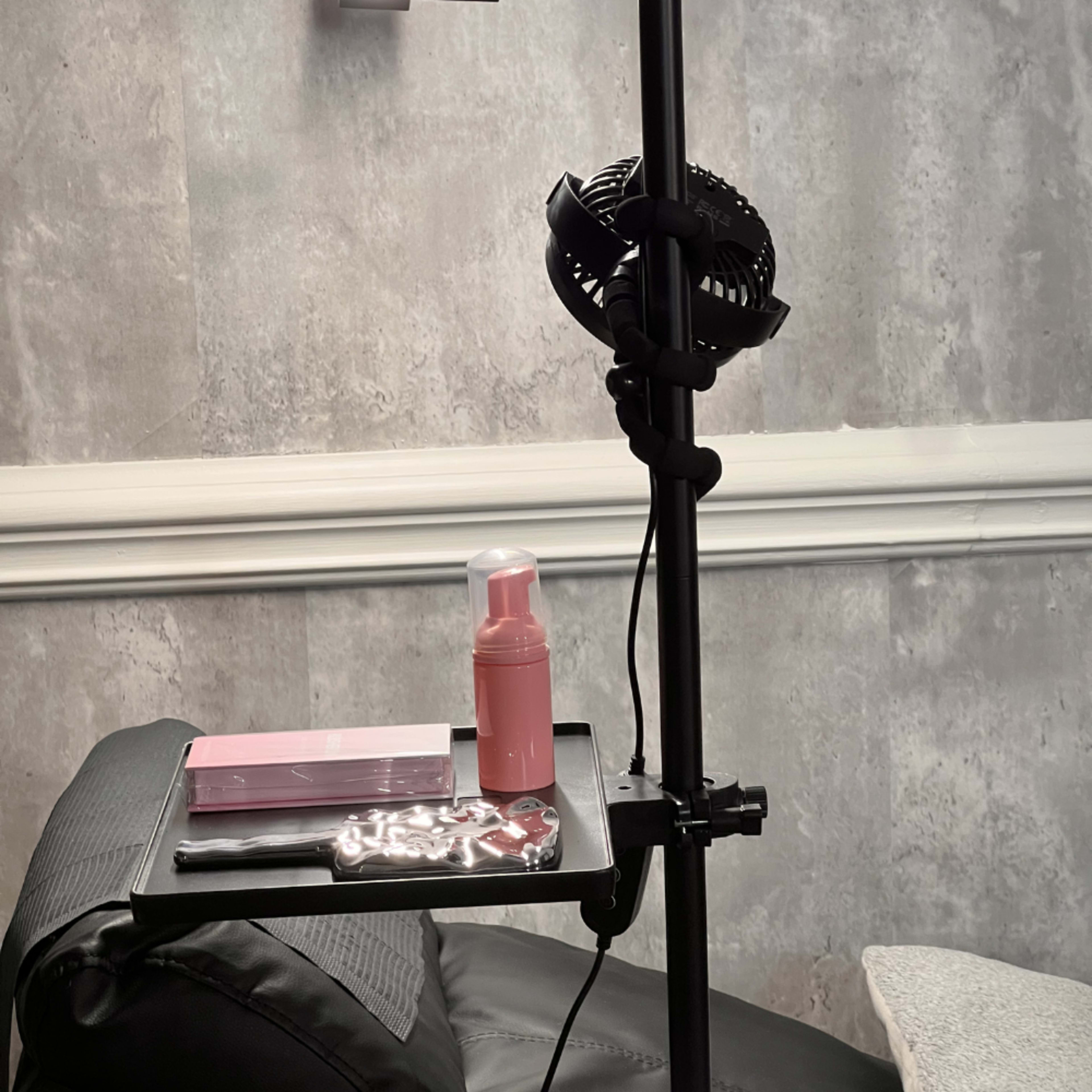 A small portable fan and various beauty products are placed on a shelf attached to a black stand next to a leather chair.