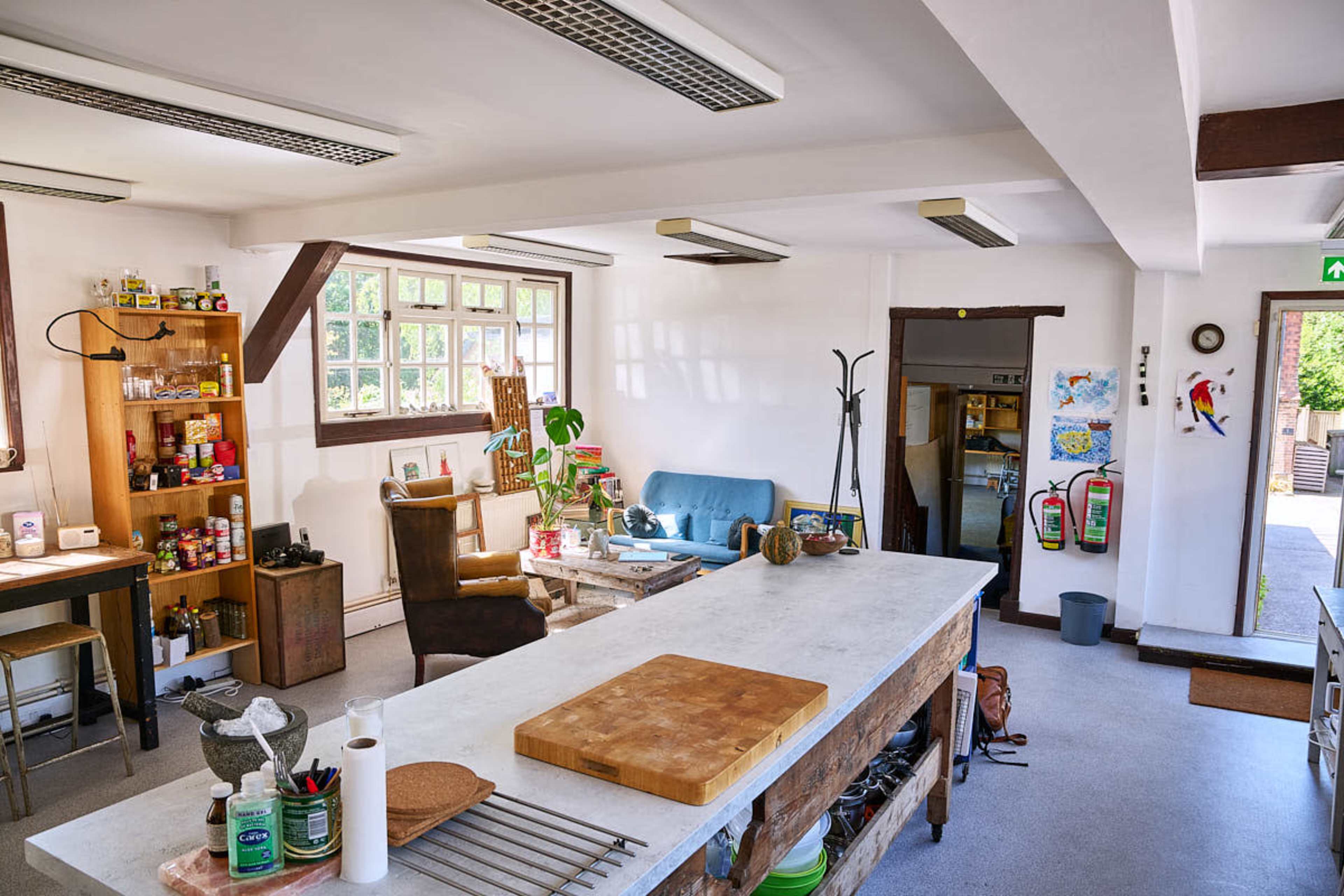 The image shows a spacious kitchen and living area with various shelves, plants, and seating arrangements, featuring a large countertop and a well-lit environment.