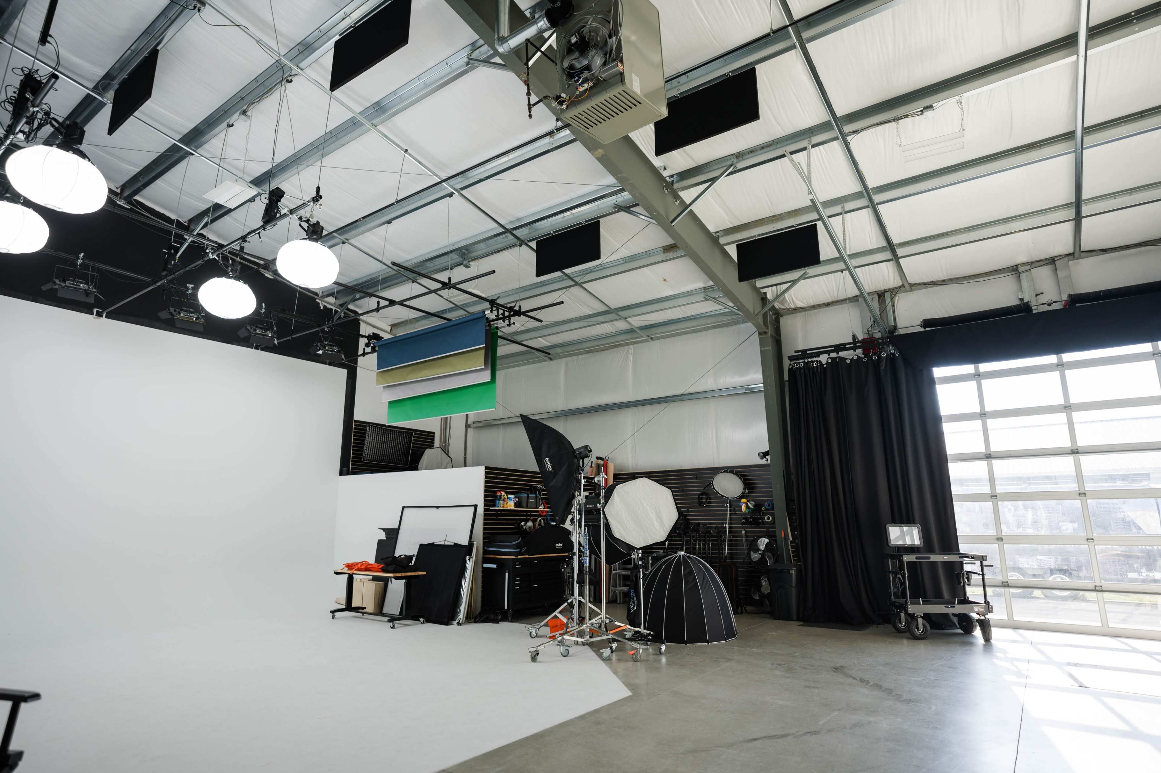 A spacious photography studio with various lighting equipment, a blank white backdrop, and a large glass door allowing natural light to enter.