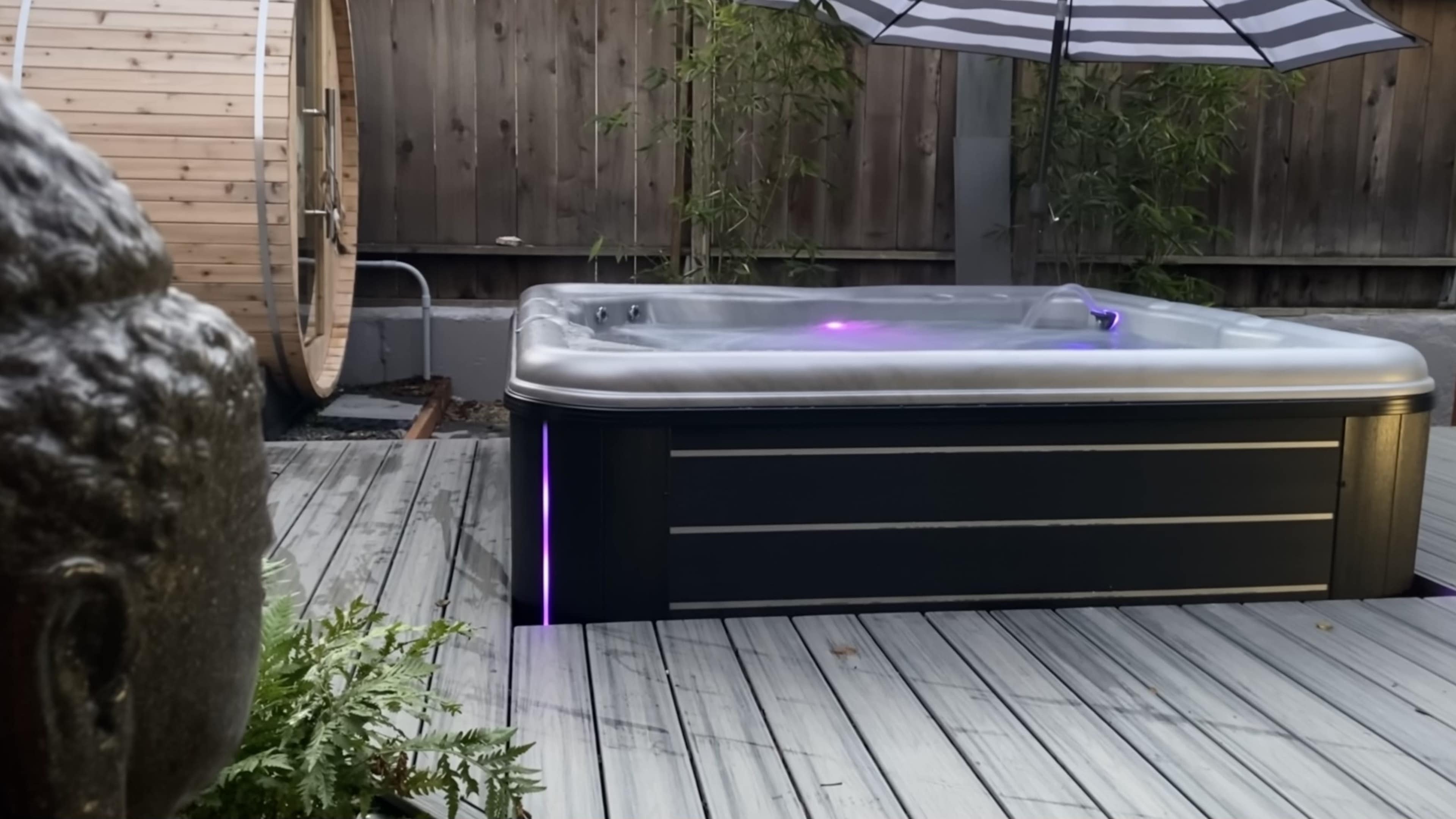 A hot tub with illuminated edges sits on a wooden deck, surrounded by greenery and a bamboo backdrop.