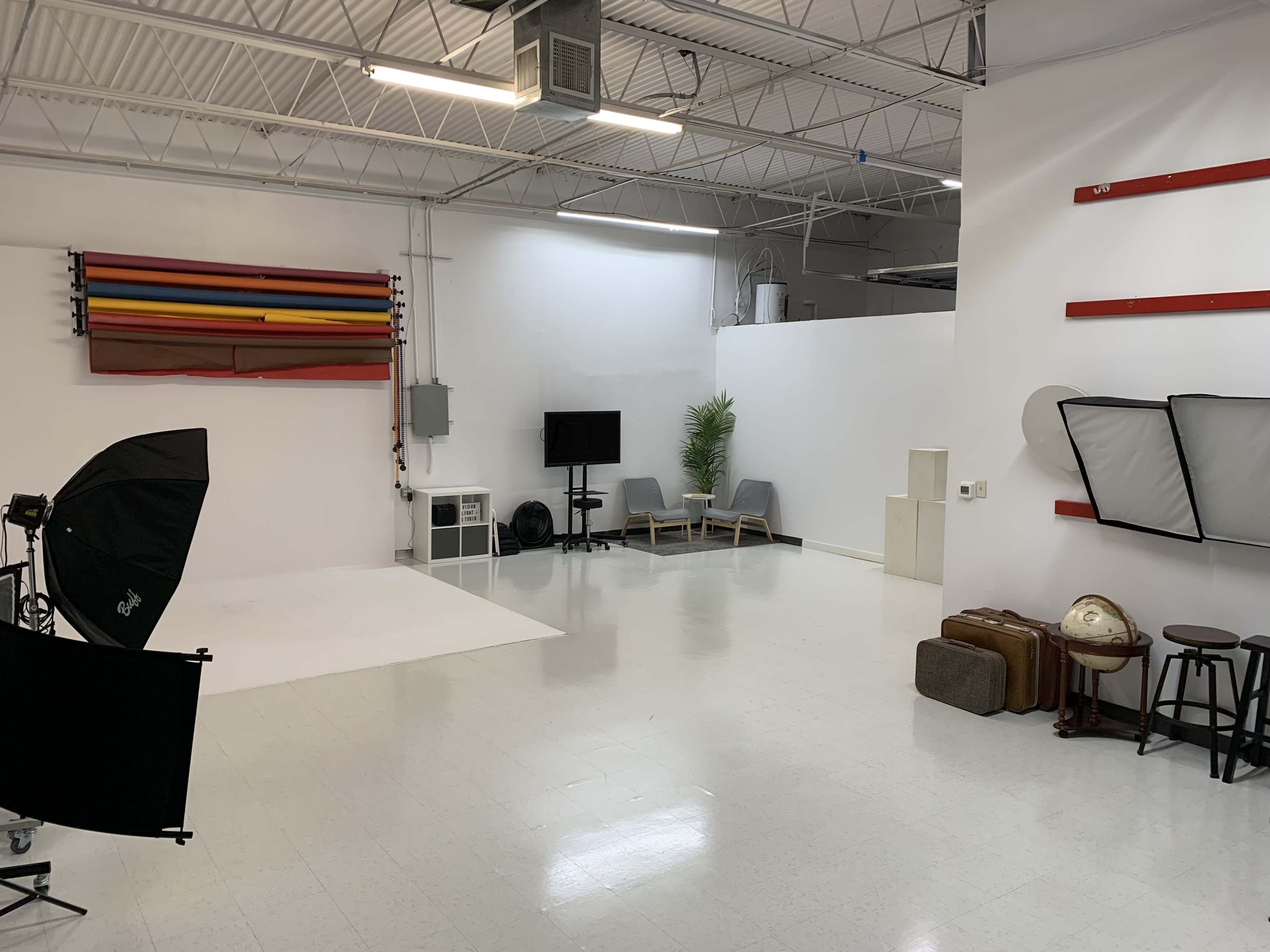A spacious studio with a white background, featuring colorful backdrop rolls, a TV, seating area, and photography equipment.