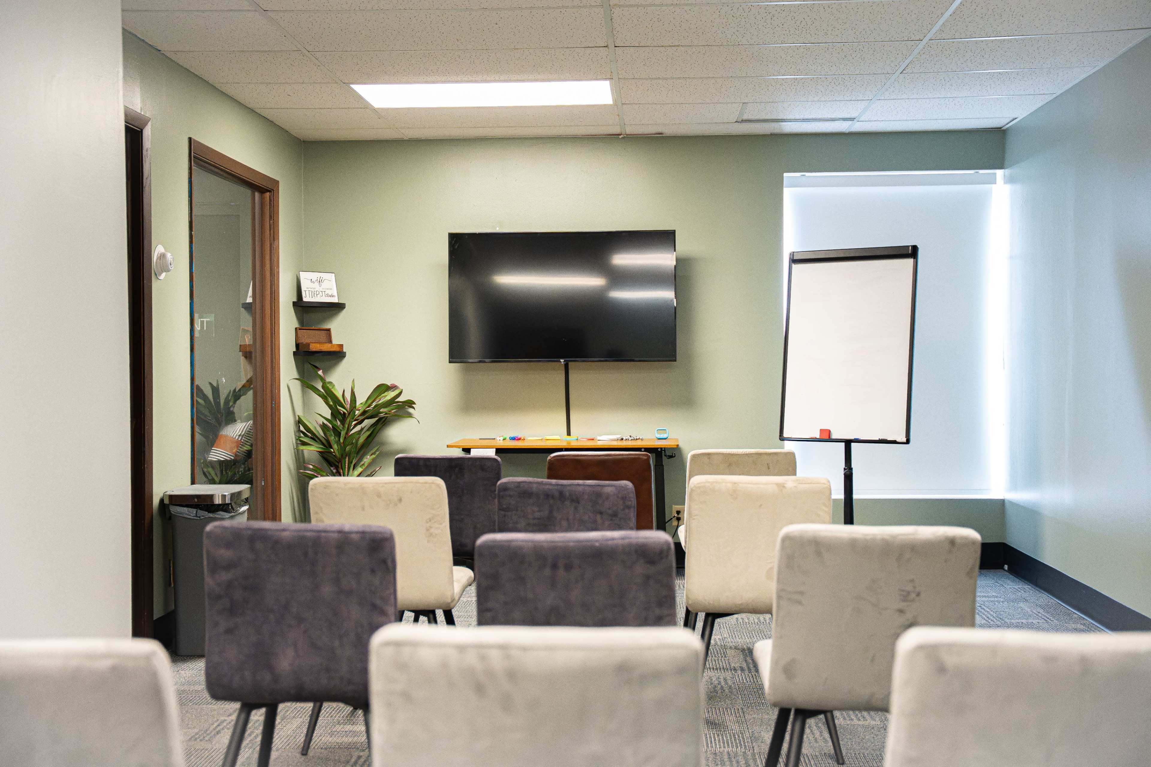 A small conference room features a television mounted on the wall, a whiteboard stand, and several chairs arranged in rows facing the screen.