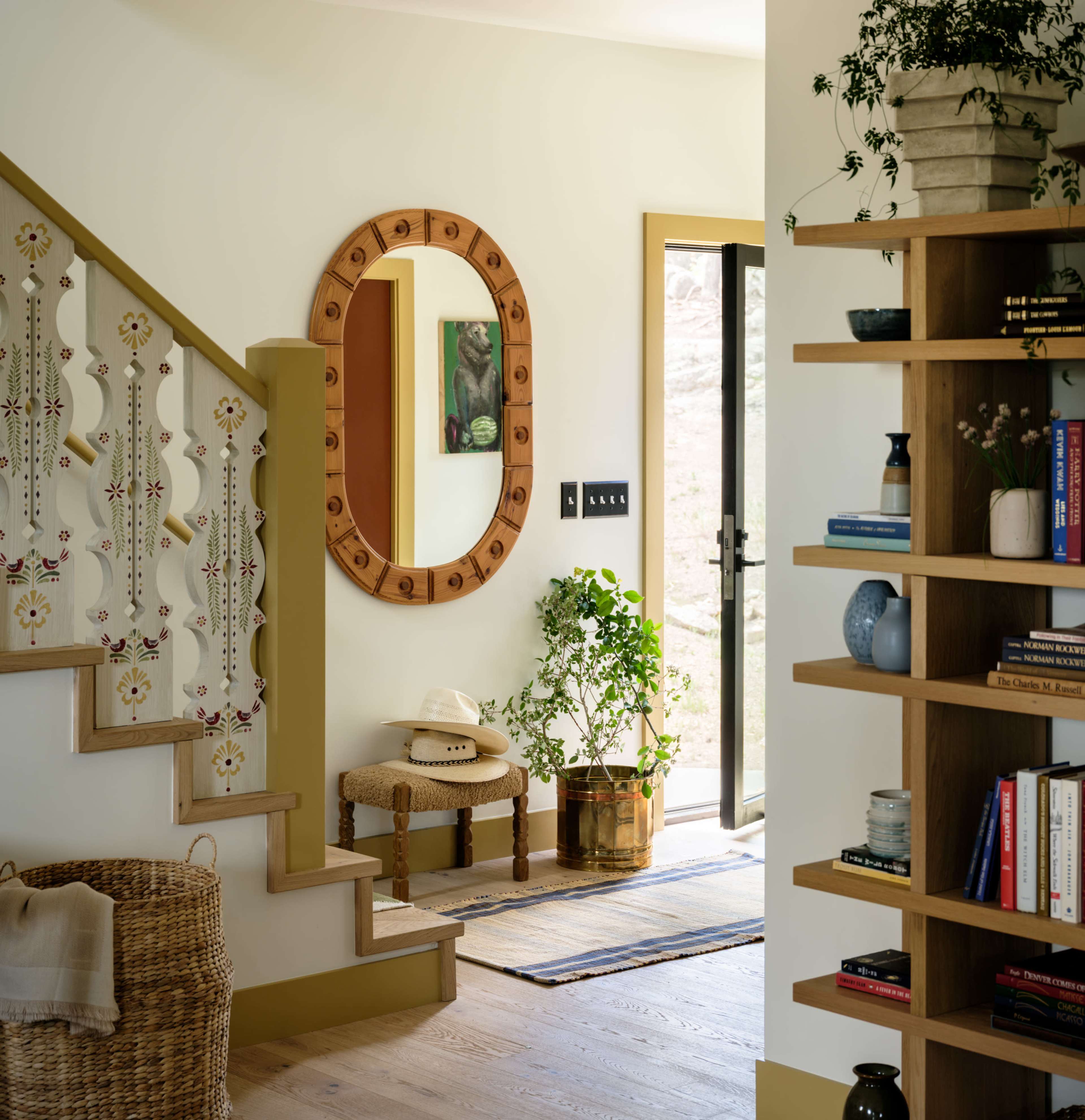 The image shows a well-lit entryway featuring a staircase with decorative railings, a round mirror, a small plant, and an open door leading outside.
