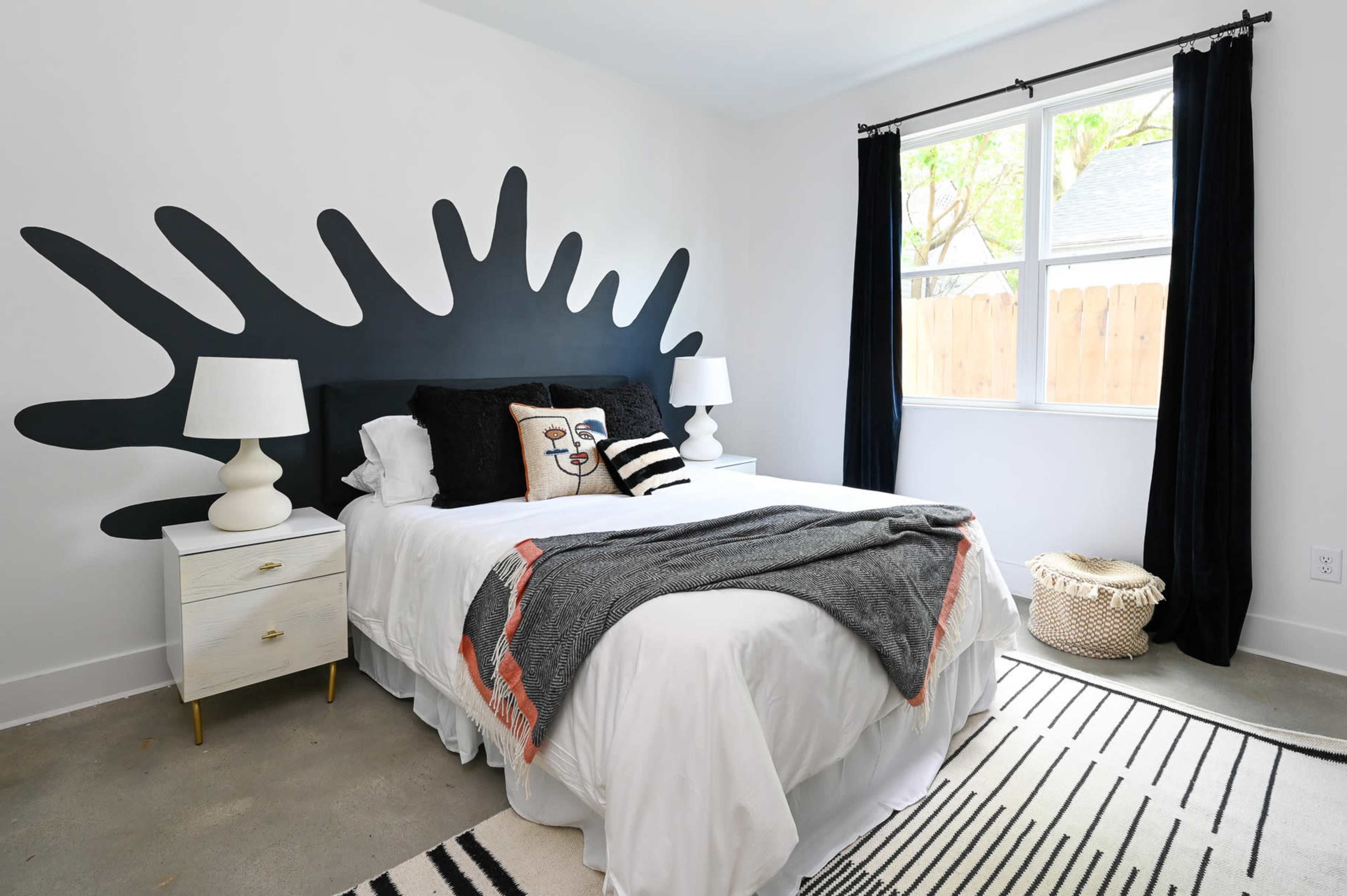 The image shows a modern bedroom with a bed, two bedside lamps, a patterned rug, and a large black abstract wall design.