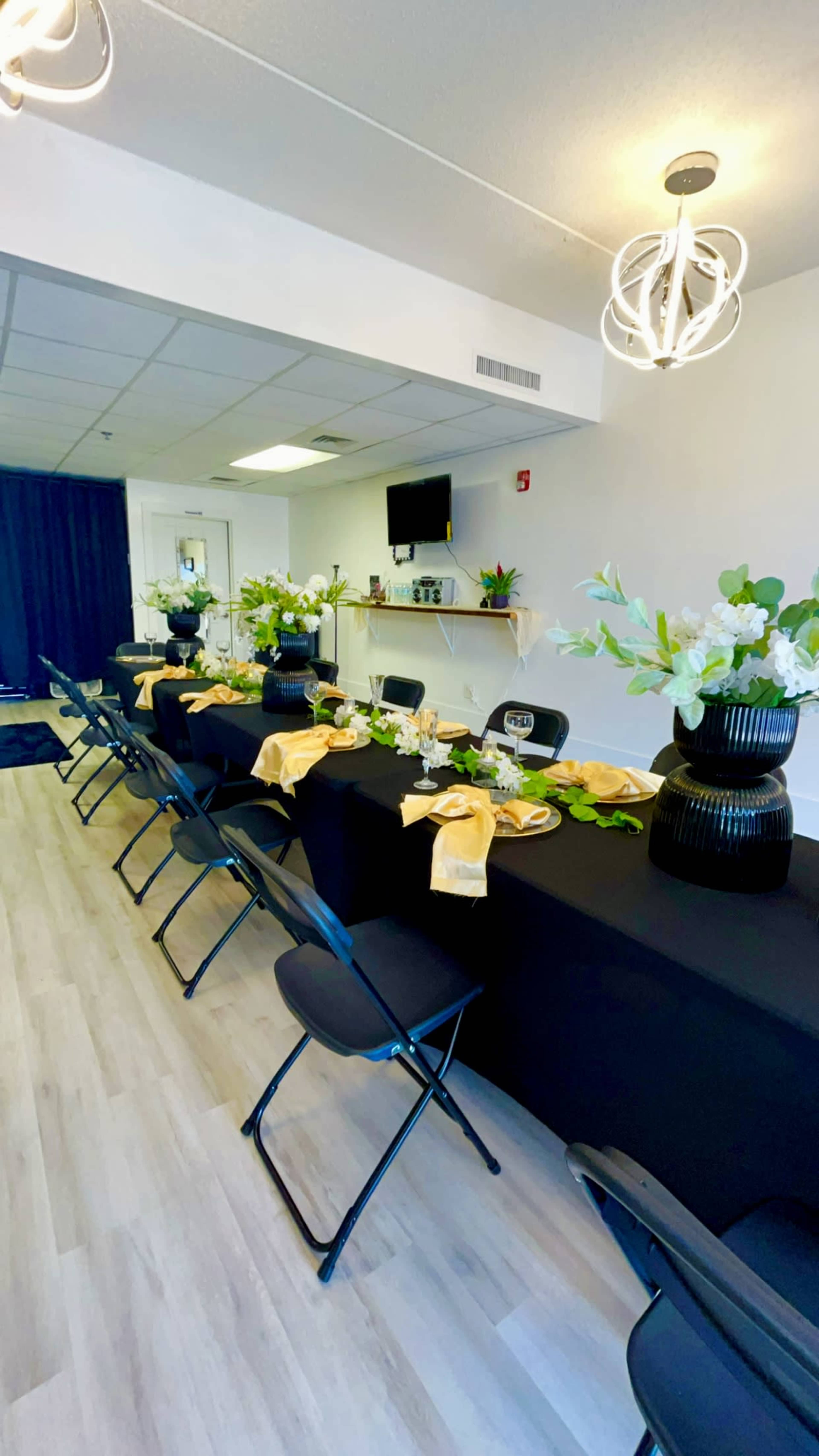 A decorated dining area features a long table set with floral centerpieces and dinnerware, surrounded by black chairs.