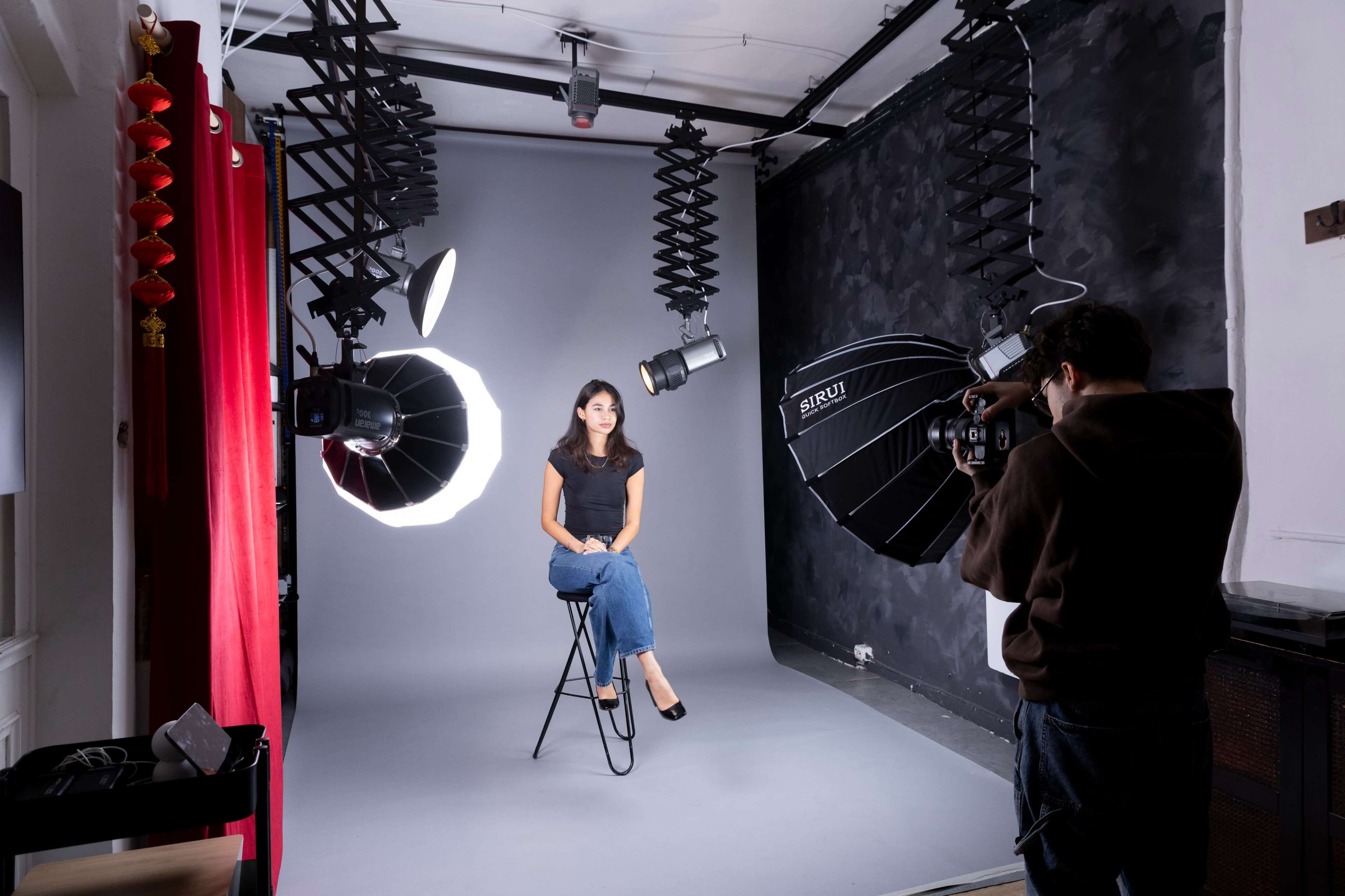 A woman sits on a stool in a photography studio, with studio lights and a gray backdrop surrounding her.
