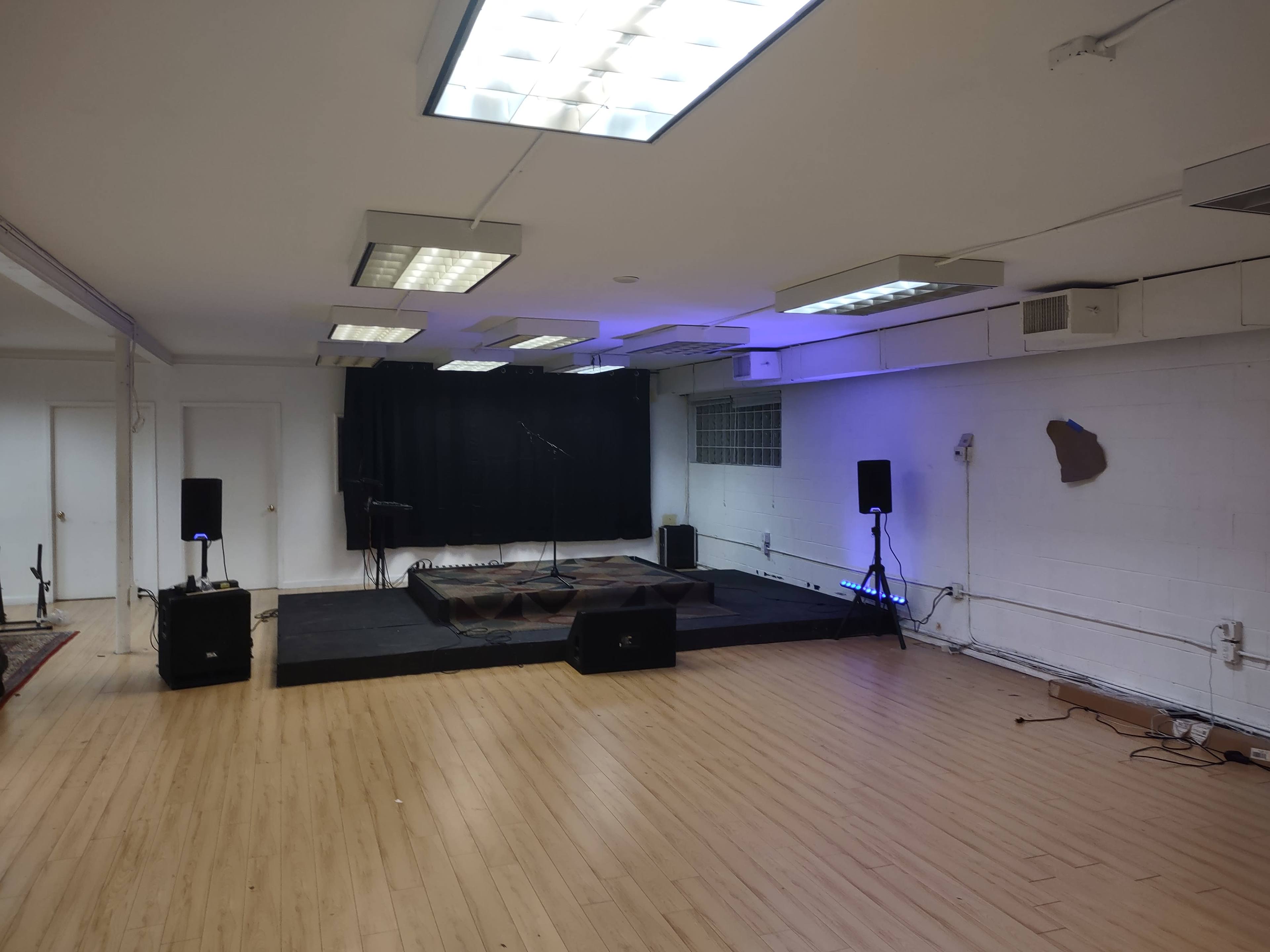 A performance space with a raised stage, microphones, and speakers set up in a room with wooden flooring and a black curtain in the background.