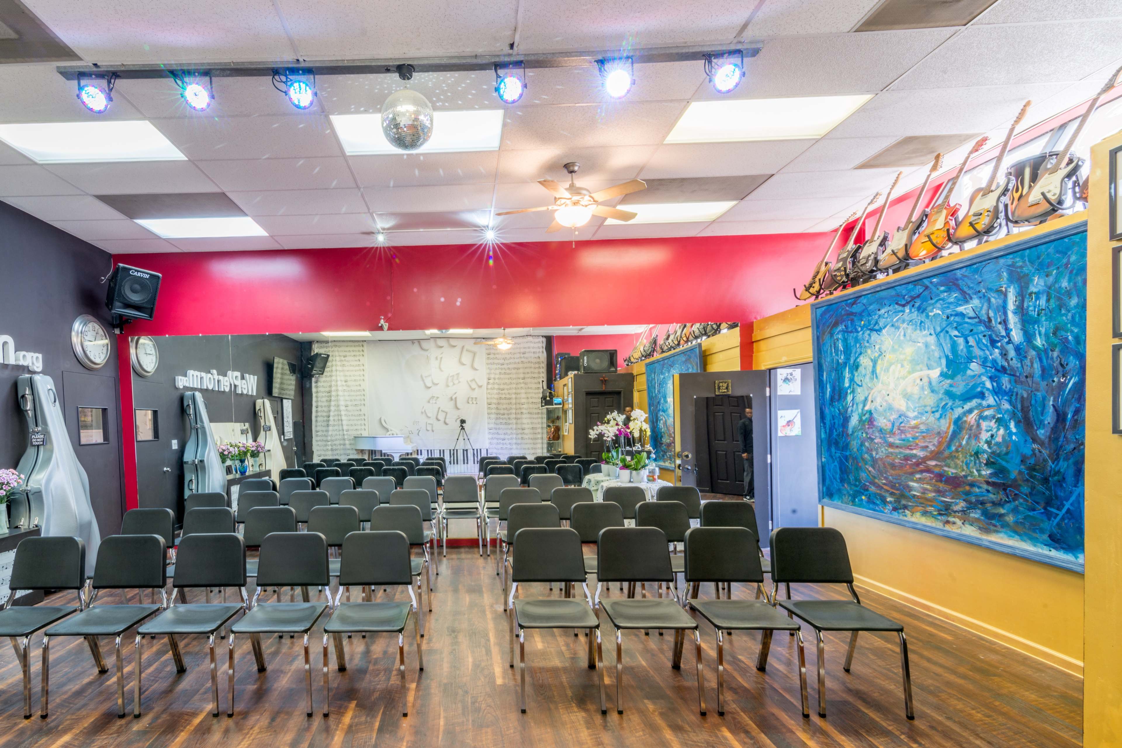 A music performance space arranged with rows of chairs facing a stage, decorated with colorful walls and guitars on display.