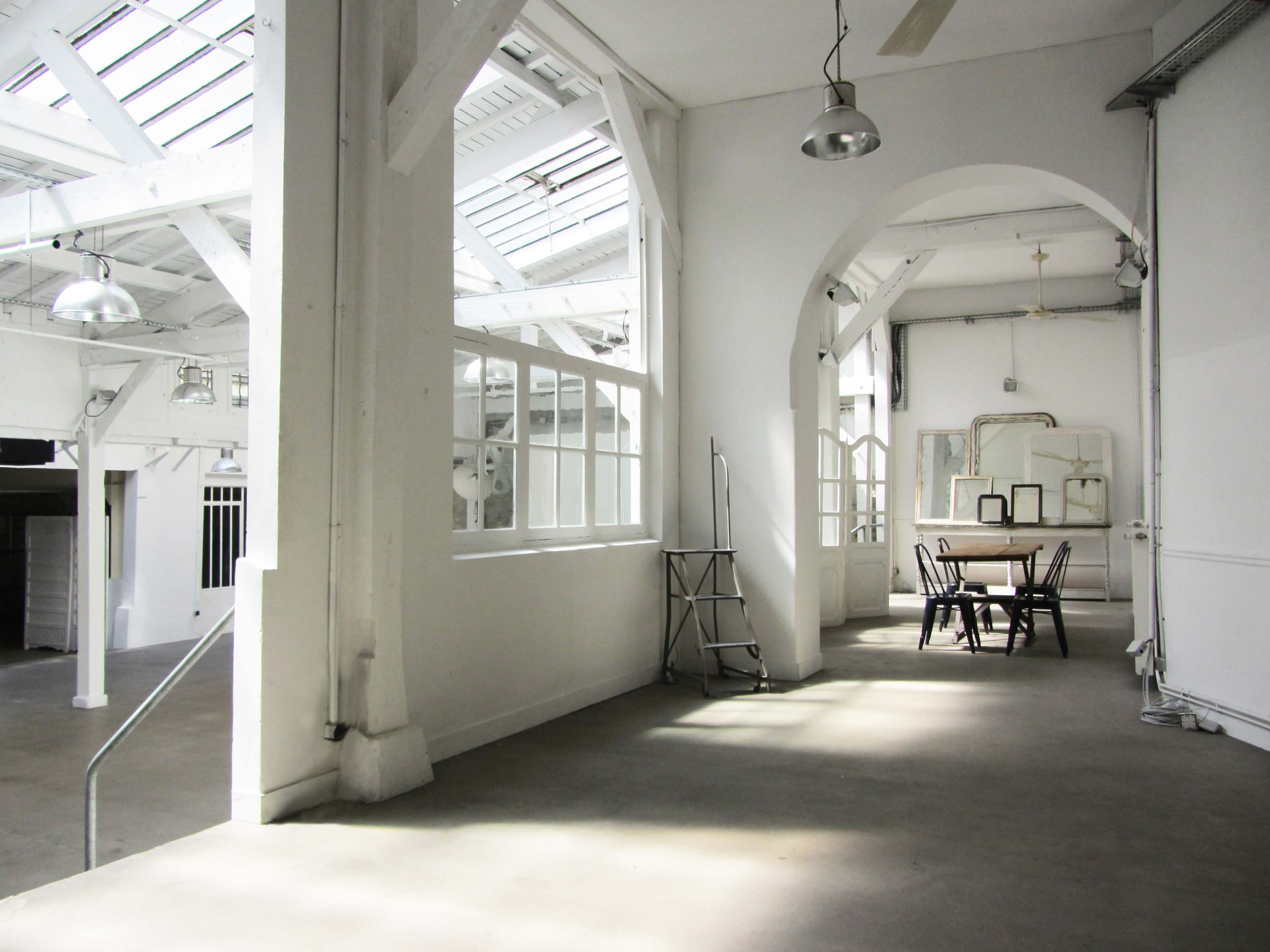 The image shows a spacious, well-lit room with large windows, white walls, and a dining table surrounded by chairs.