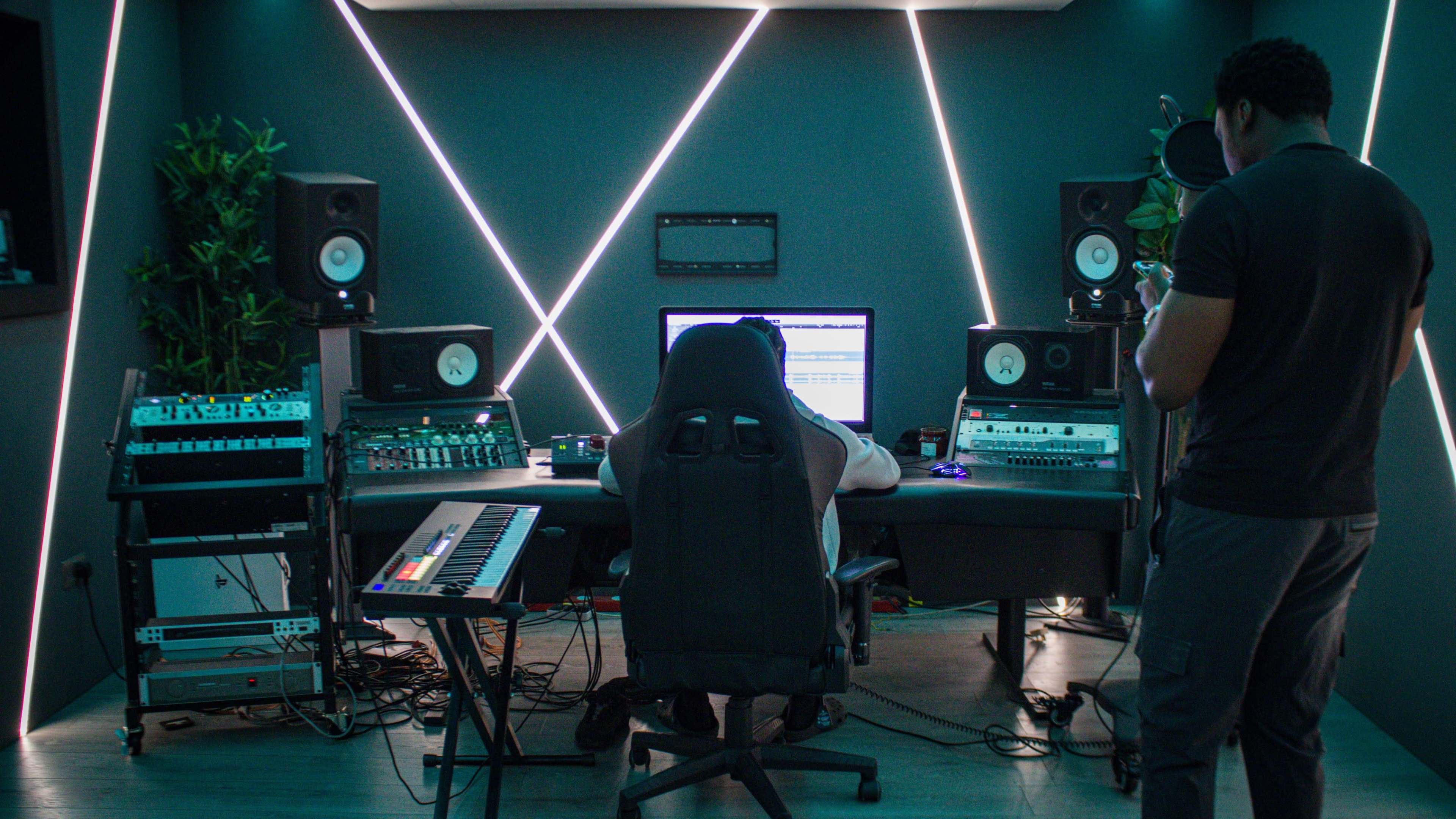 A music production studio features a person seated at a desk with computer equipment and audio gear, while another individual stands nearby, looking at a phone.