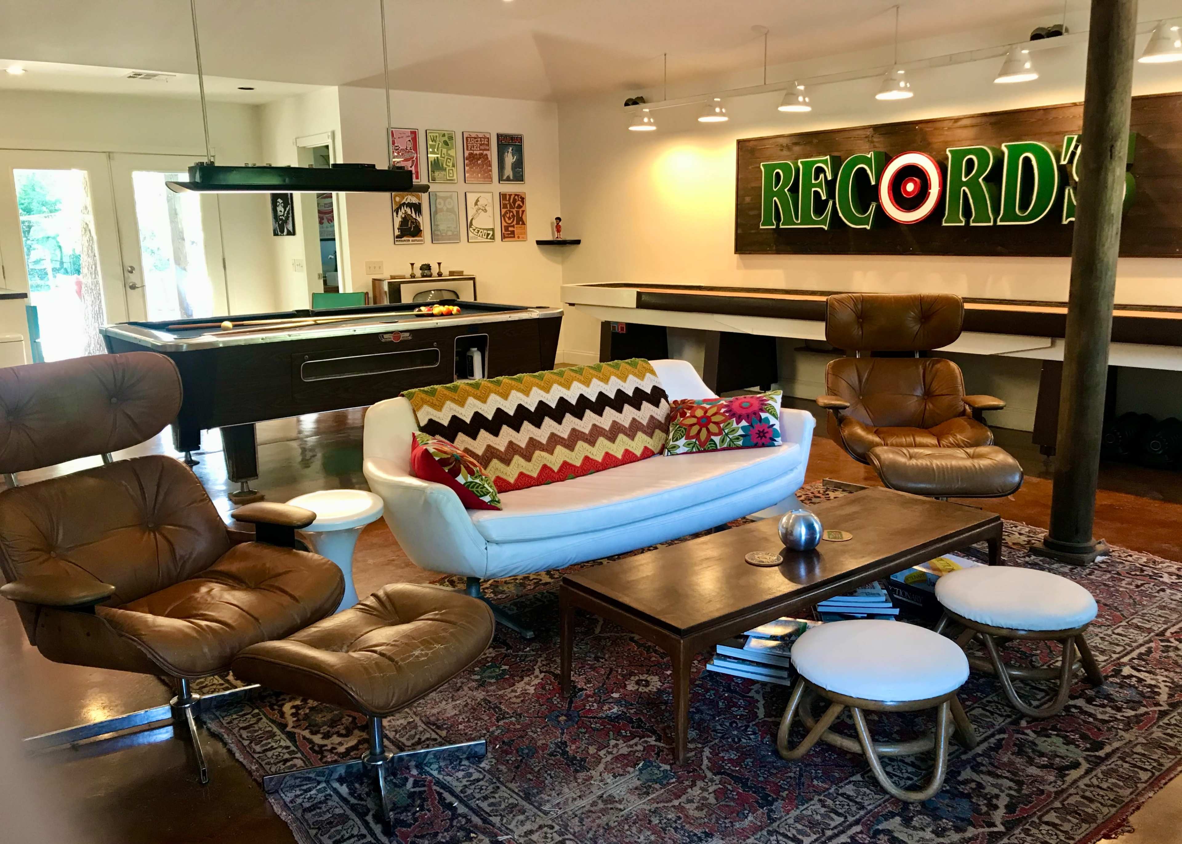 A modern living room with a white sofa, two brown lounge chairs, a wooden coffee table, and a wall featuring colorful record-themed artwork and a neon sign that says "RECORDS."