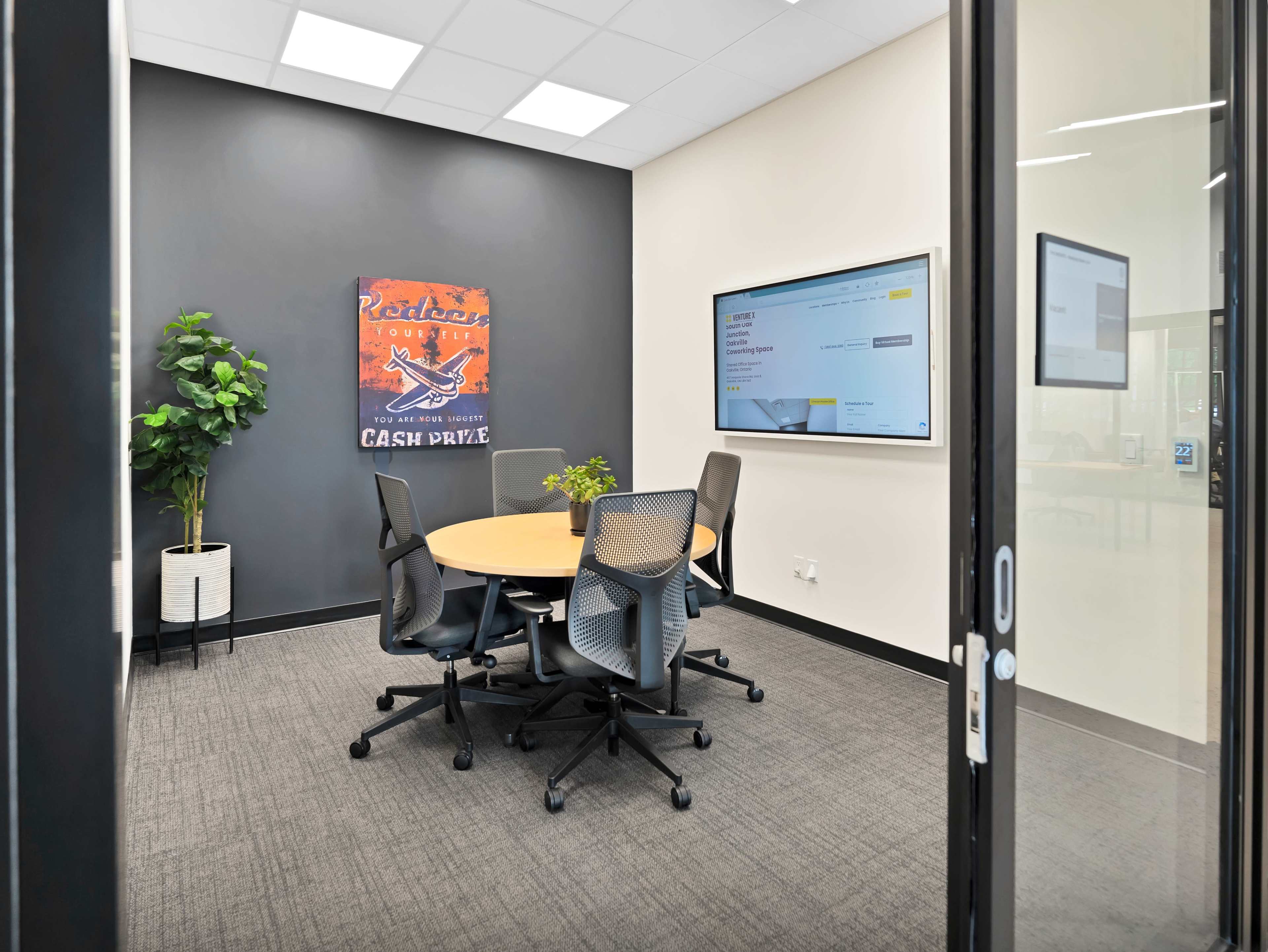 A well-lit meeting room features a round table surrounded by four chairs, a potted plant, and a large screen displaying digital content on one wall.