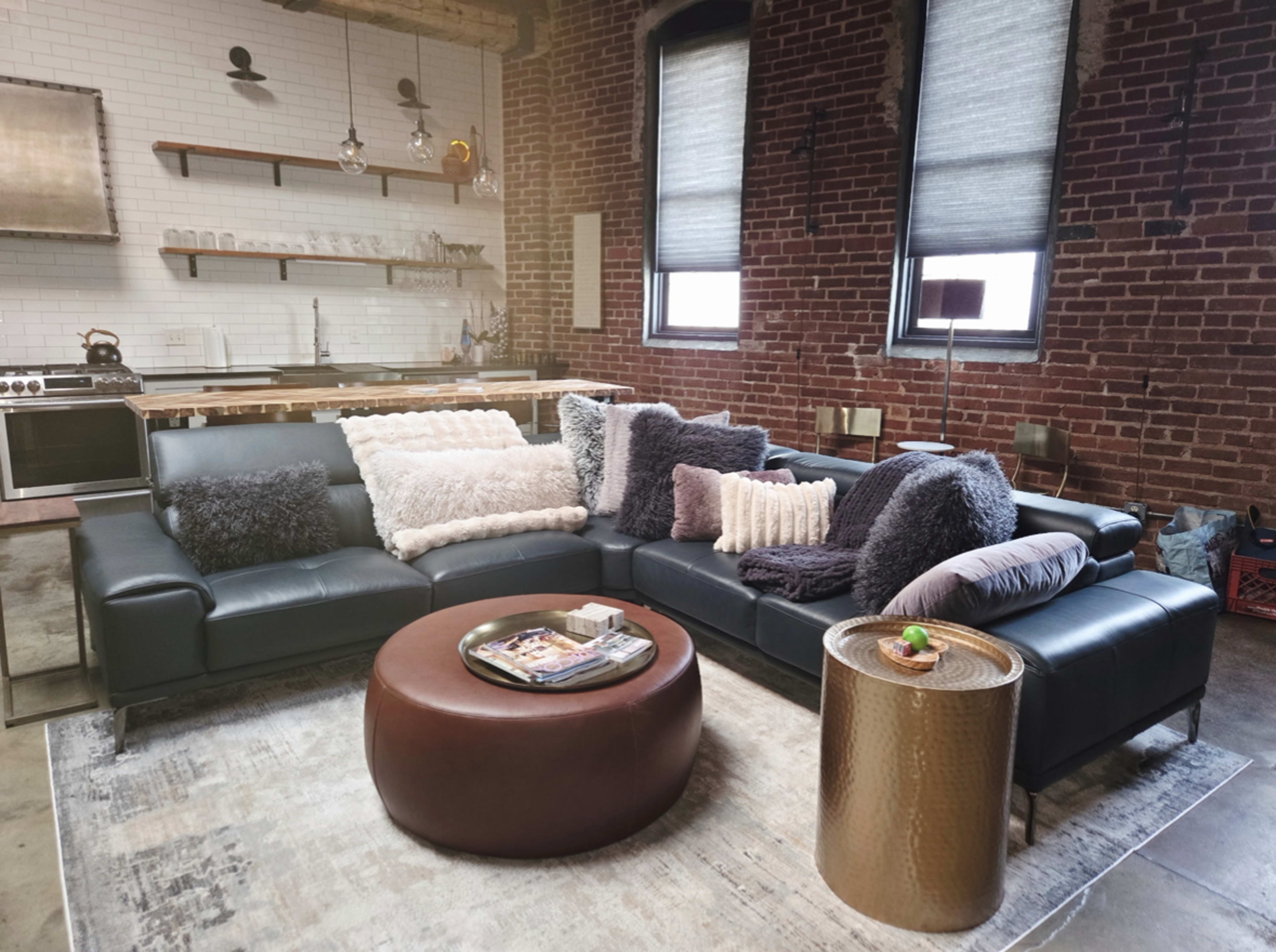 The image shows a modern living room featuring a large sectional sofa adorned with various textured pillows, a round coffee table, and a kitchen area with exposed brick walls and minimalist decor in the background.