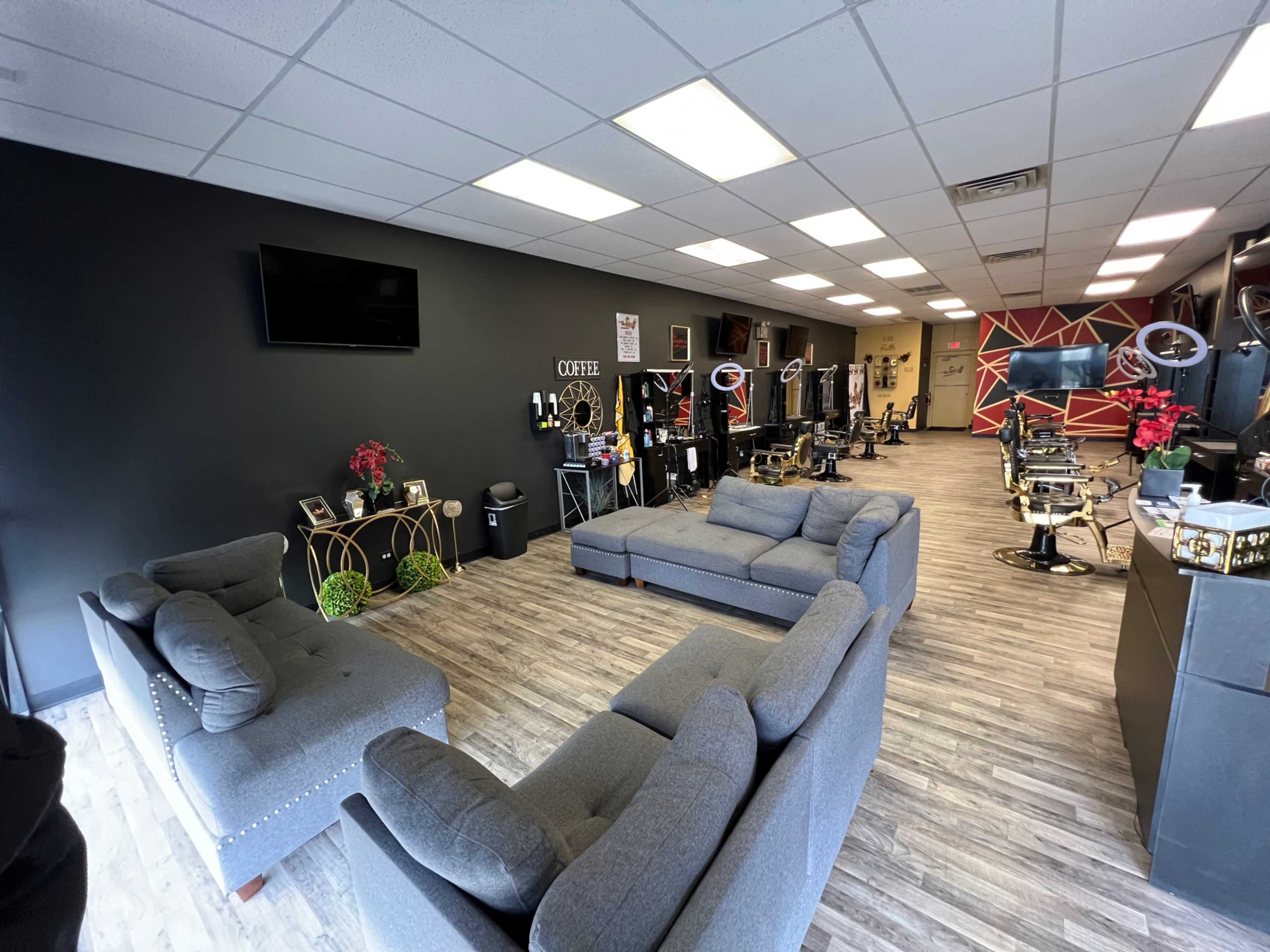 A barber shop interior with several styling stations, two gray sofas, and a waiting area against a dark wall.