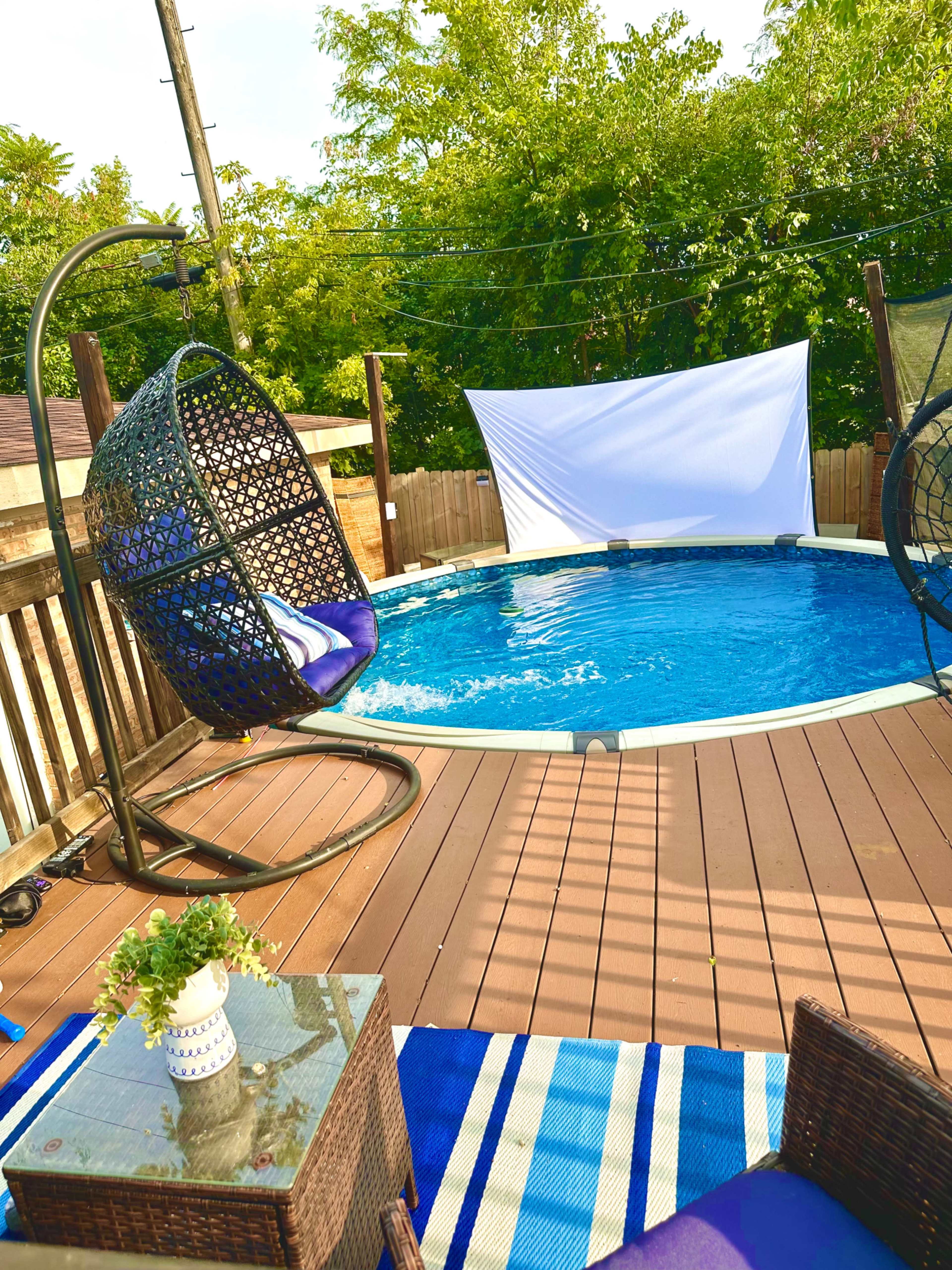 A round above-ground pool surrounded by a wooden deck with two hanging chairs and a white screen in the background.