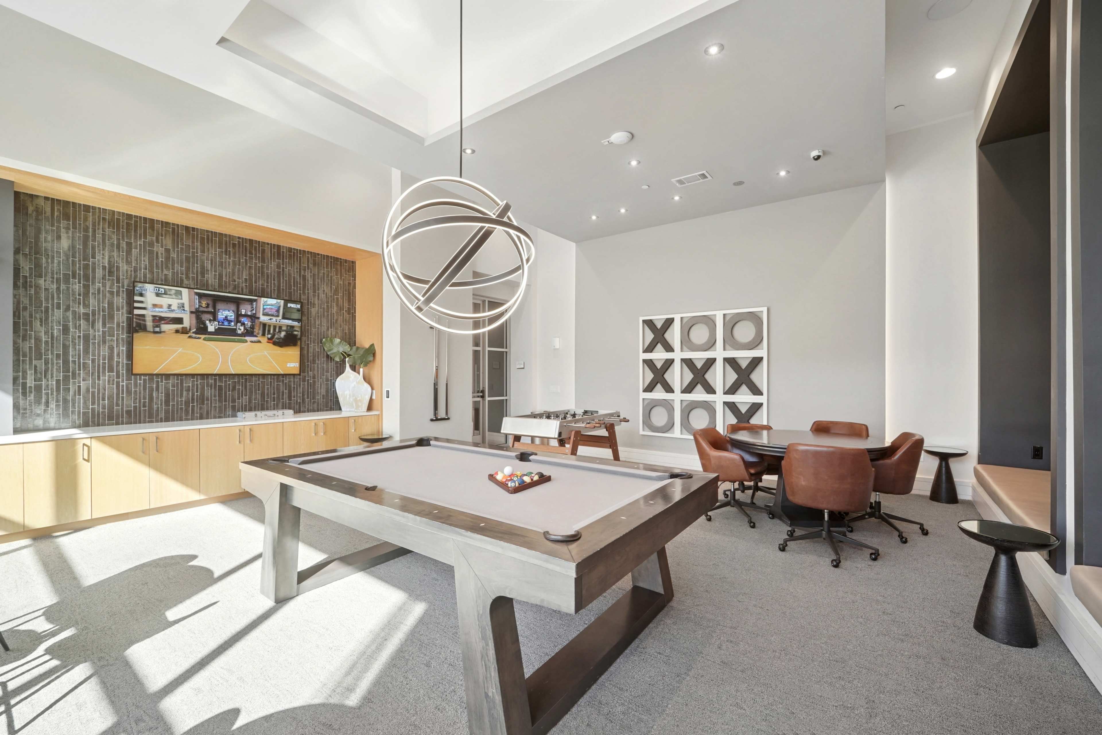 The image shows a modern recreational room featuring a pool table, a wall-mounted television, a dining area with a round table and chairs, and contemporary decor.