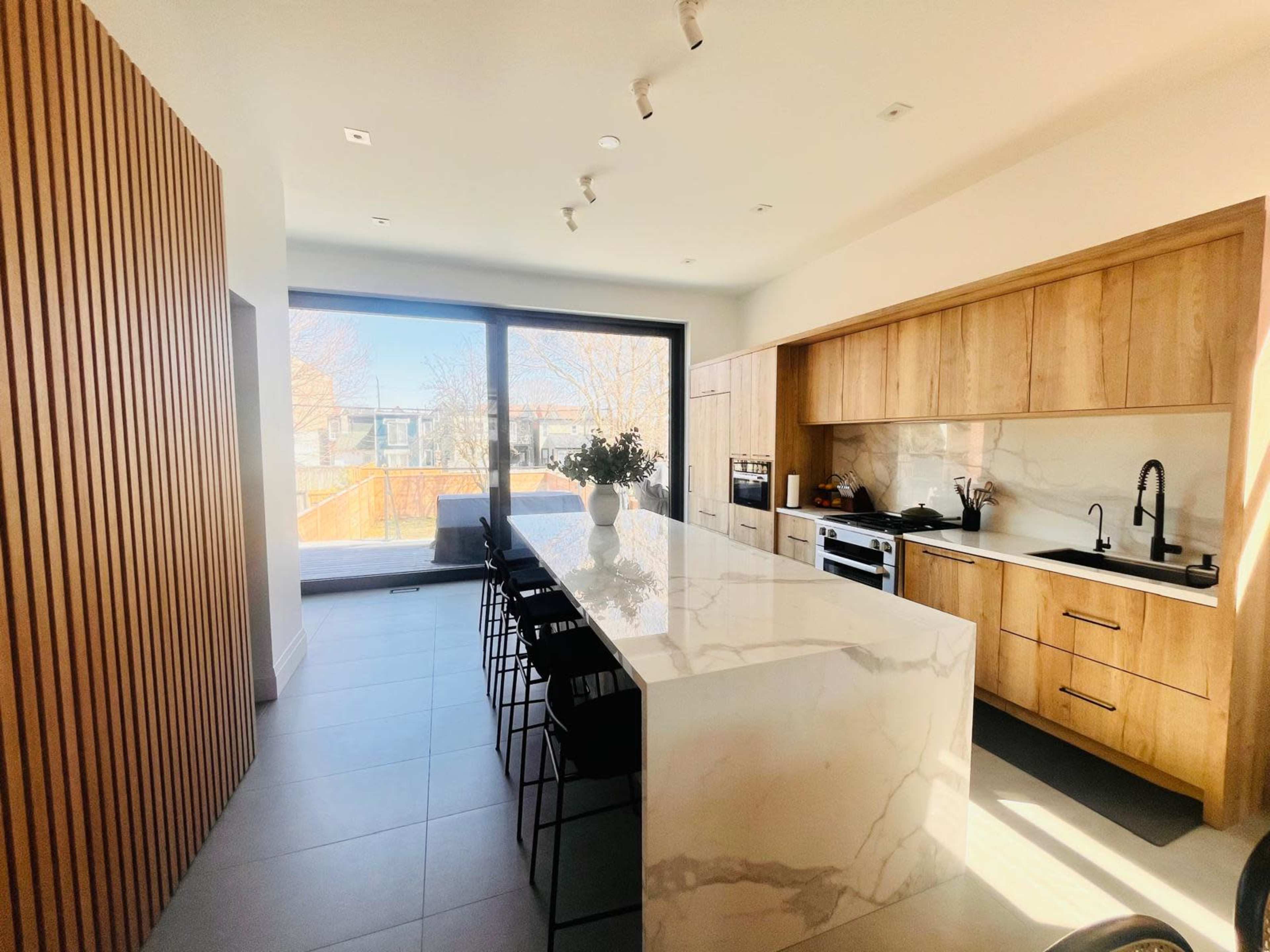 The image shows a modern kitchen with a large marble island, wooden cabinetry, and a large window overlooking an outdoor area.