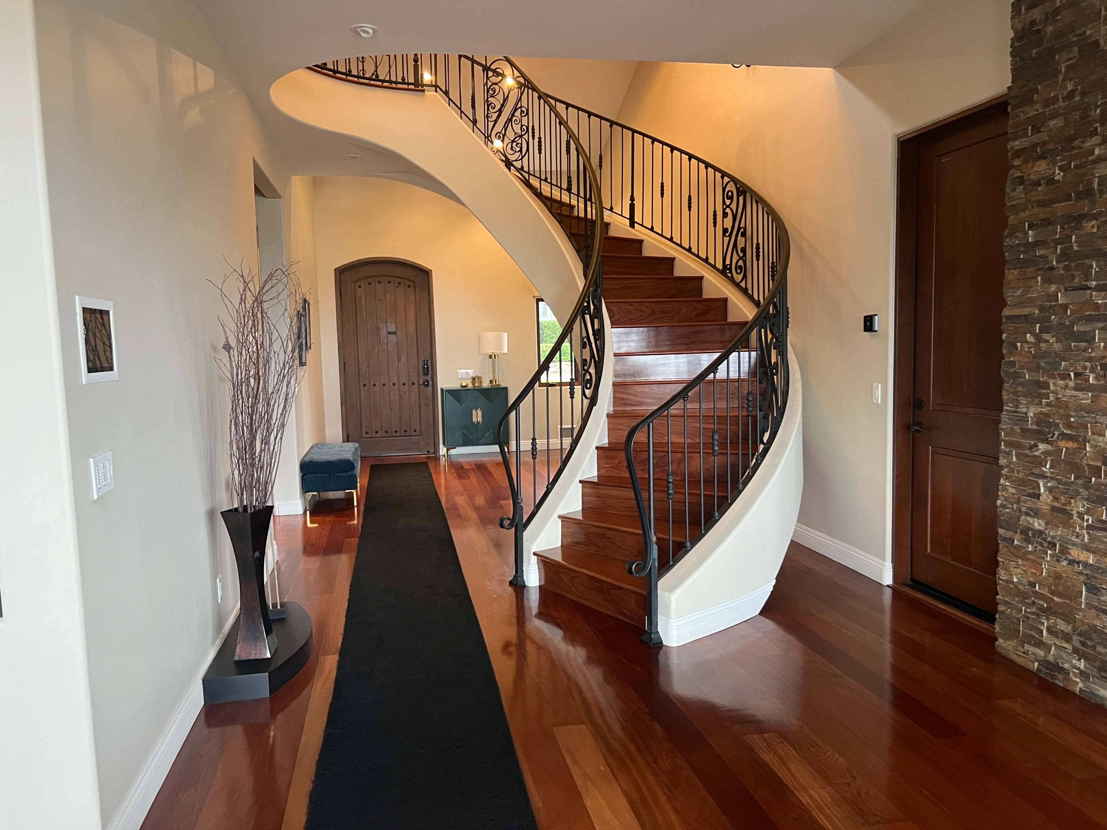 A curved wooden staircase with wrought iron railings ascends from a spacious entryway featuring a dark runner rug and decorative accents.