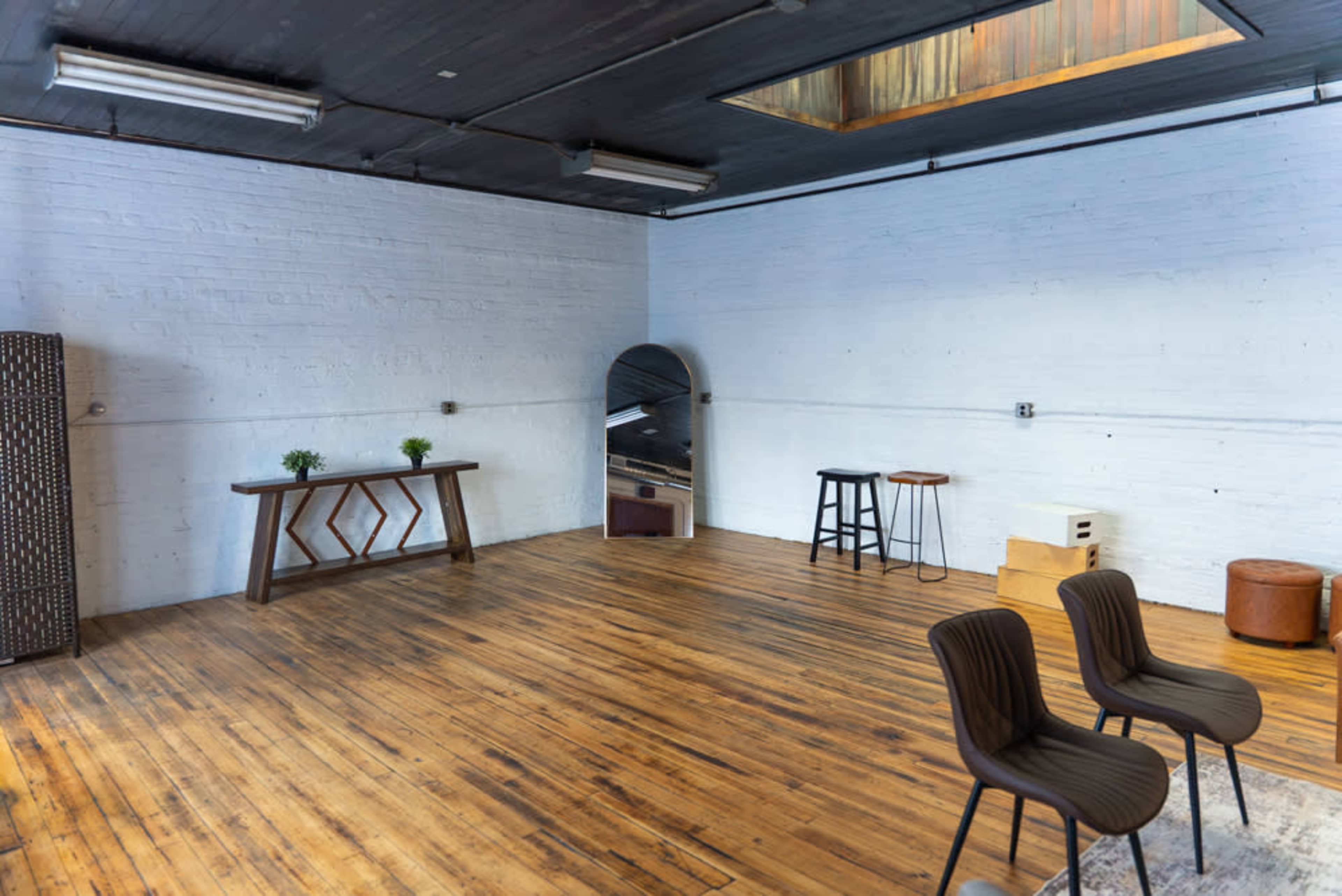The image shows a spacious, empty room with wooden floors, a large mirror, two black chairs, and various furniture pieces against the white brick walls.