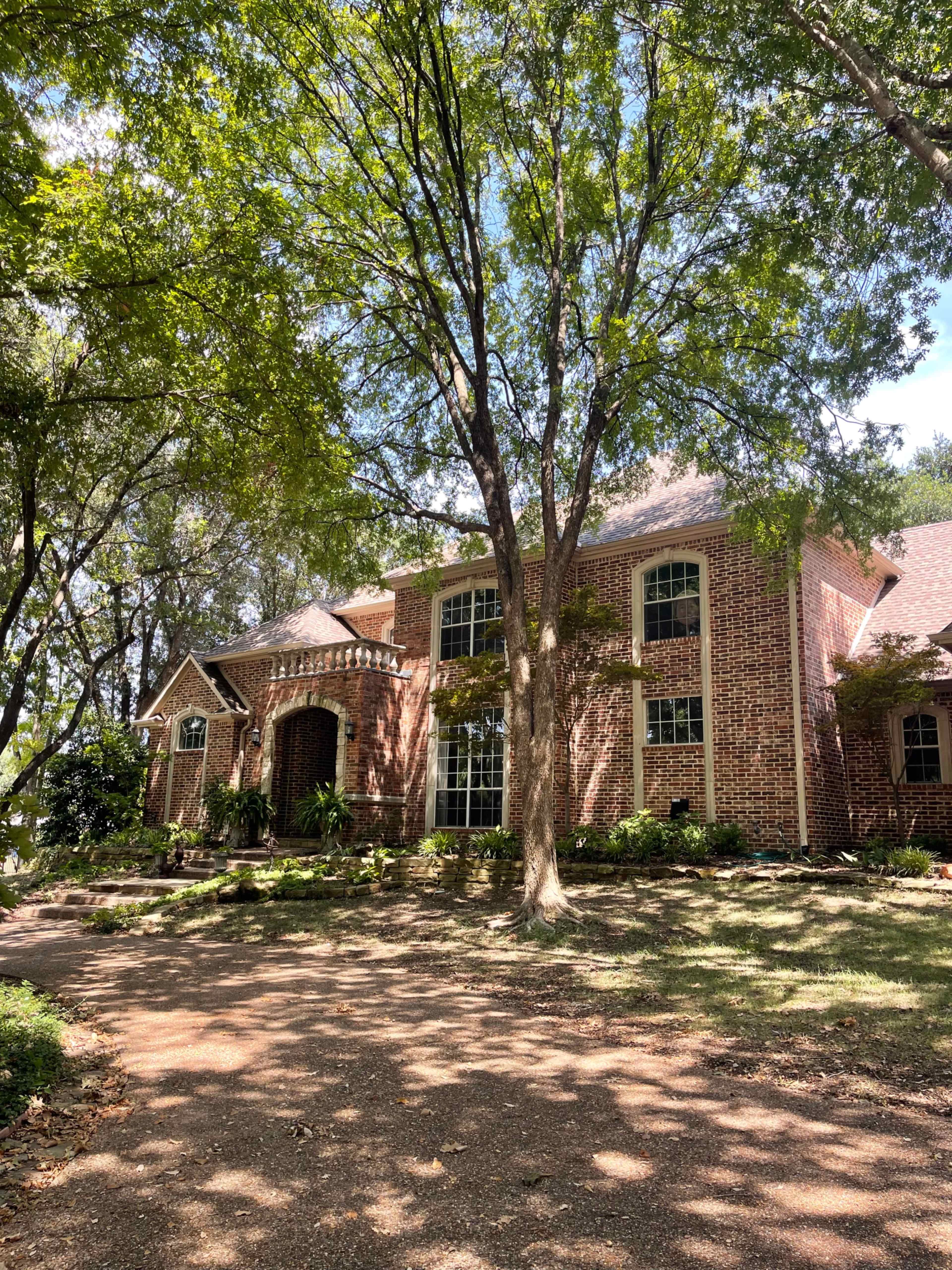 A two-story brick house is surrounded by trees and greenery, with a gravel driveway leading up to it.