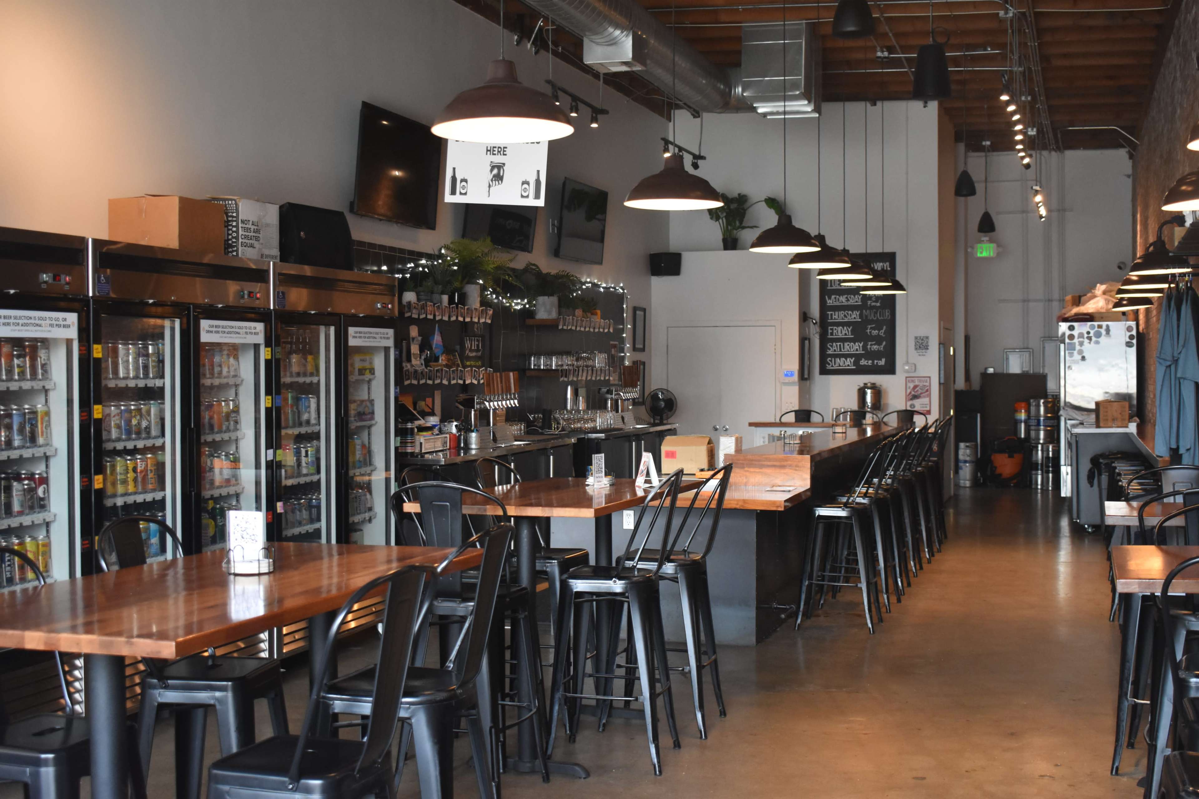 The image shows the interior of a modern bar with a long countertop, black stools, and a row of refrigerators filled with drinks along one wall.