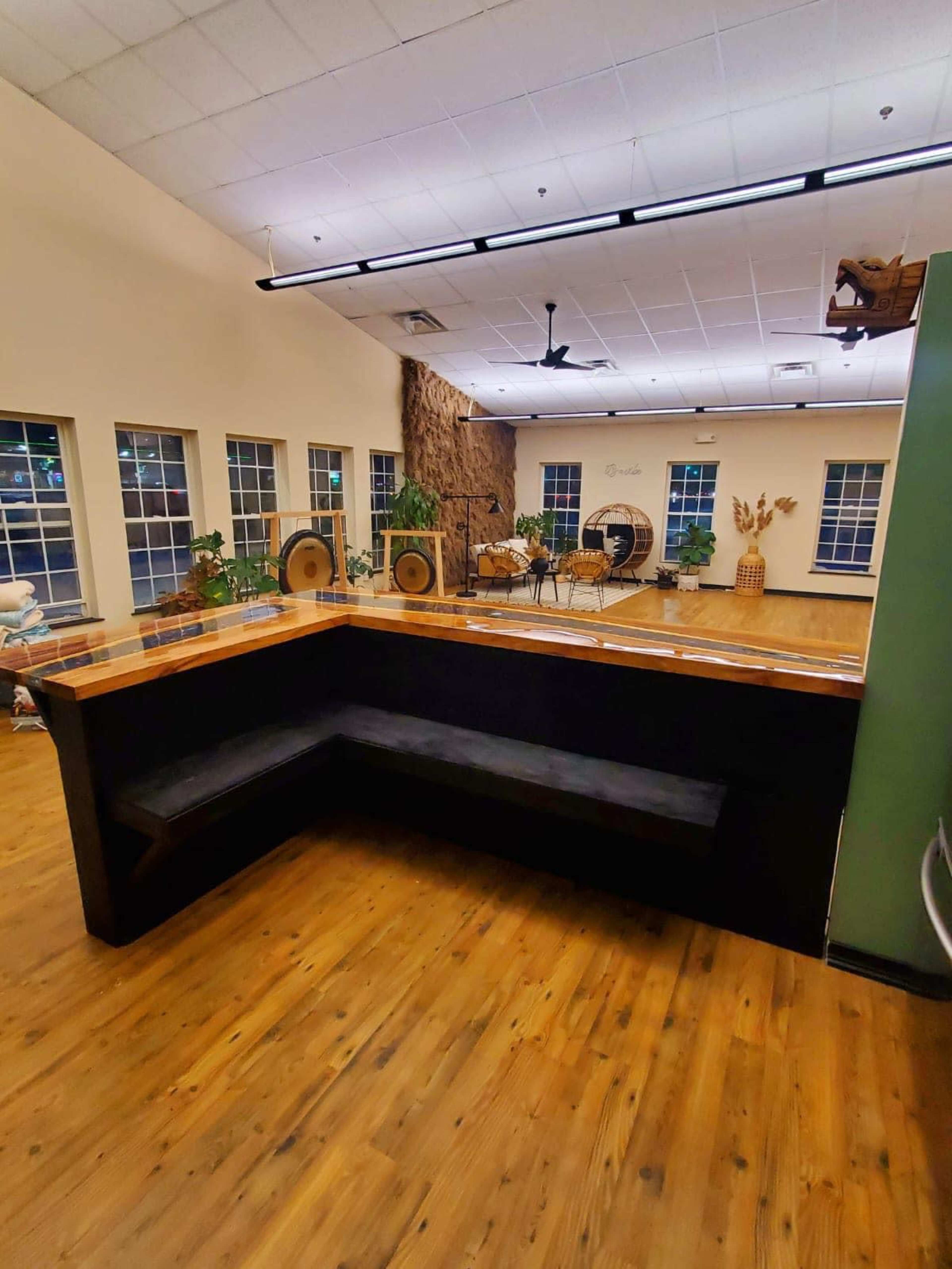 The image shows a spacious room with wooden flooring, featuring a black seating area against the wall and large windows allowing natural light.