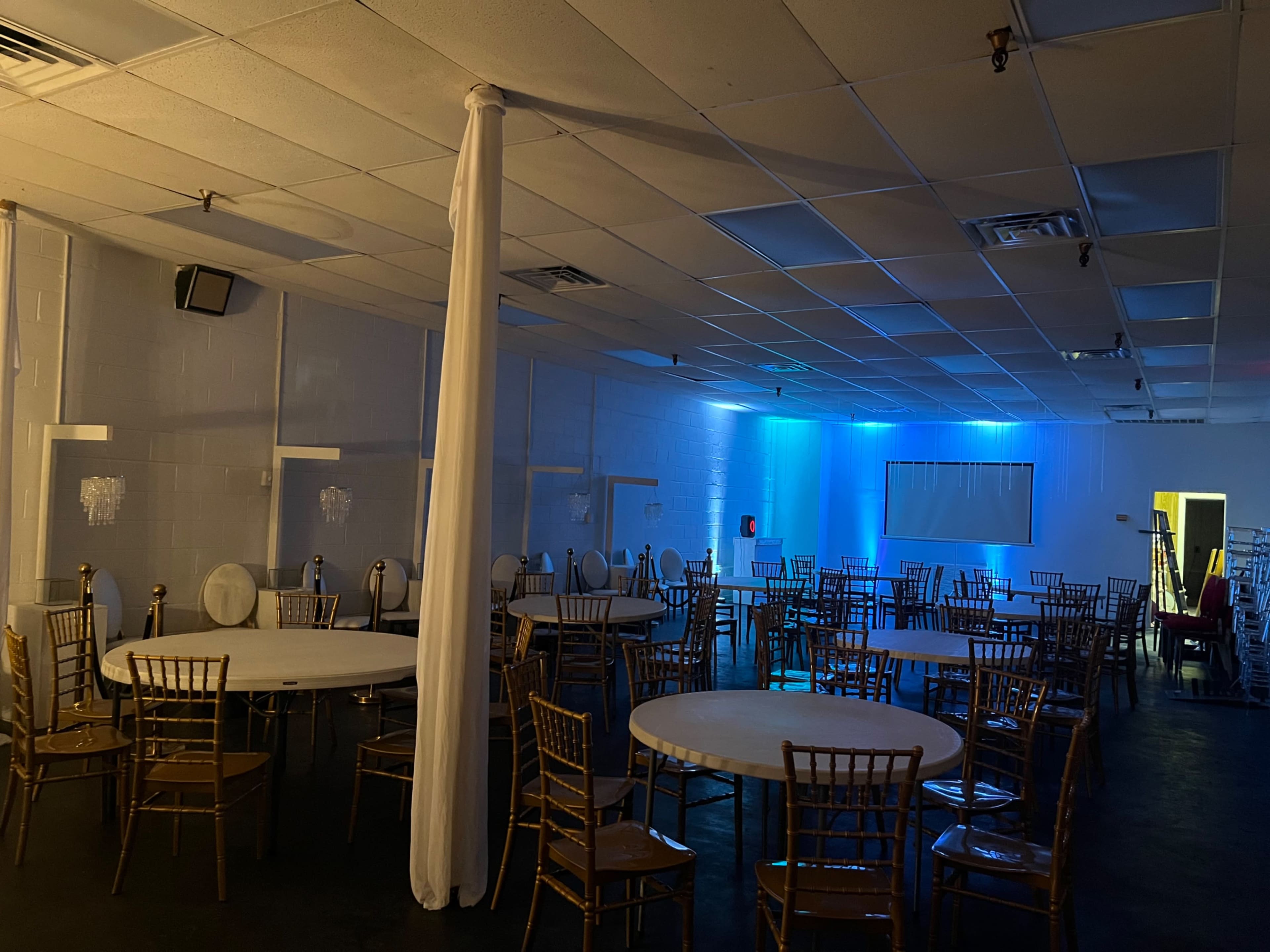 A sparsely furnished event space with round tables and gold chairs, illuminated by blue lighting in one corner.