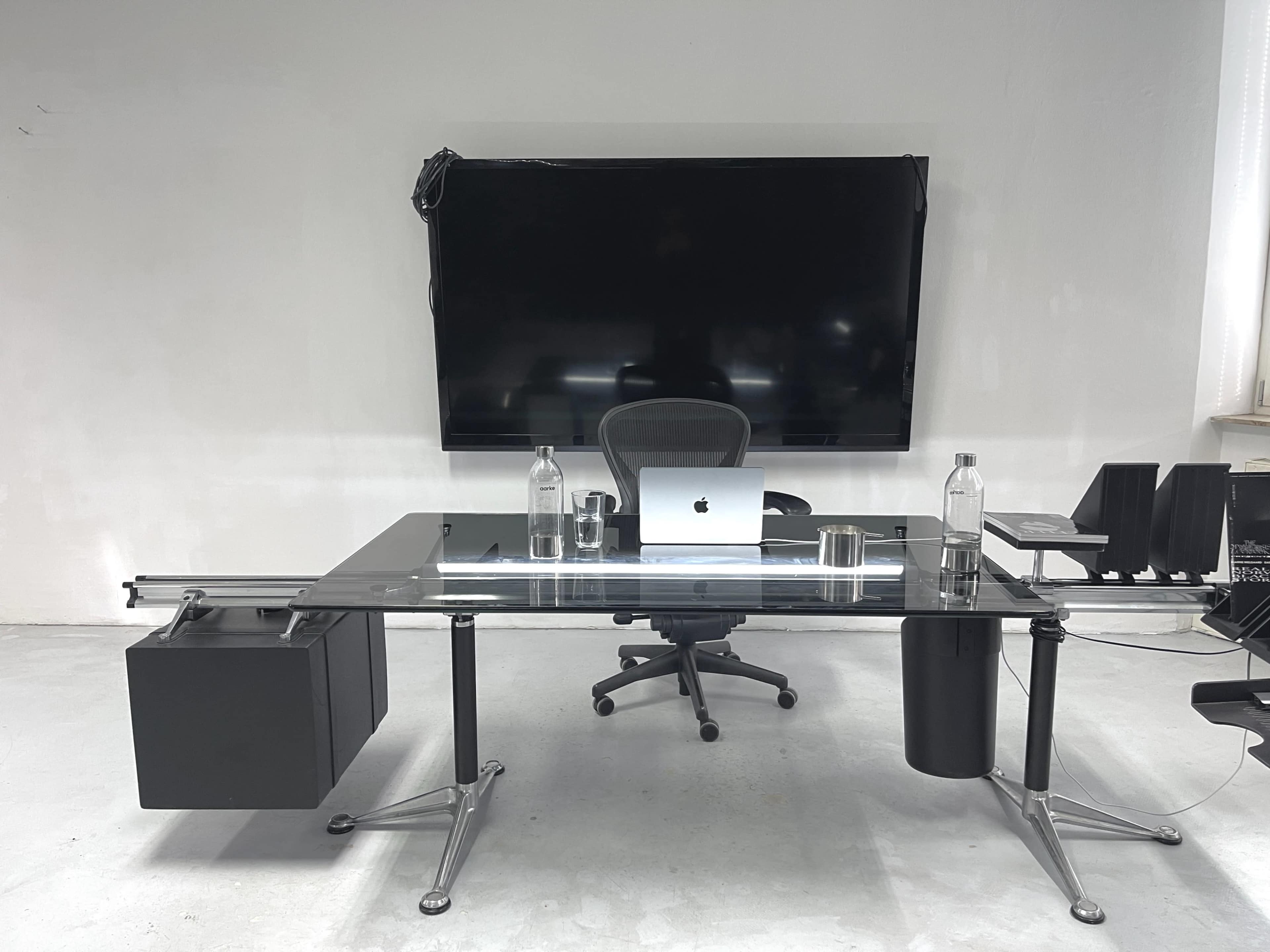 A modern office setup features a glass desk with an office chair, a laptop, and water bottles, positioned in front of a large flat-screen TV.