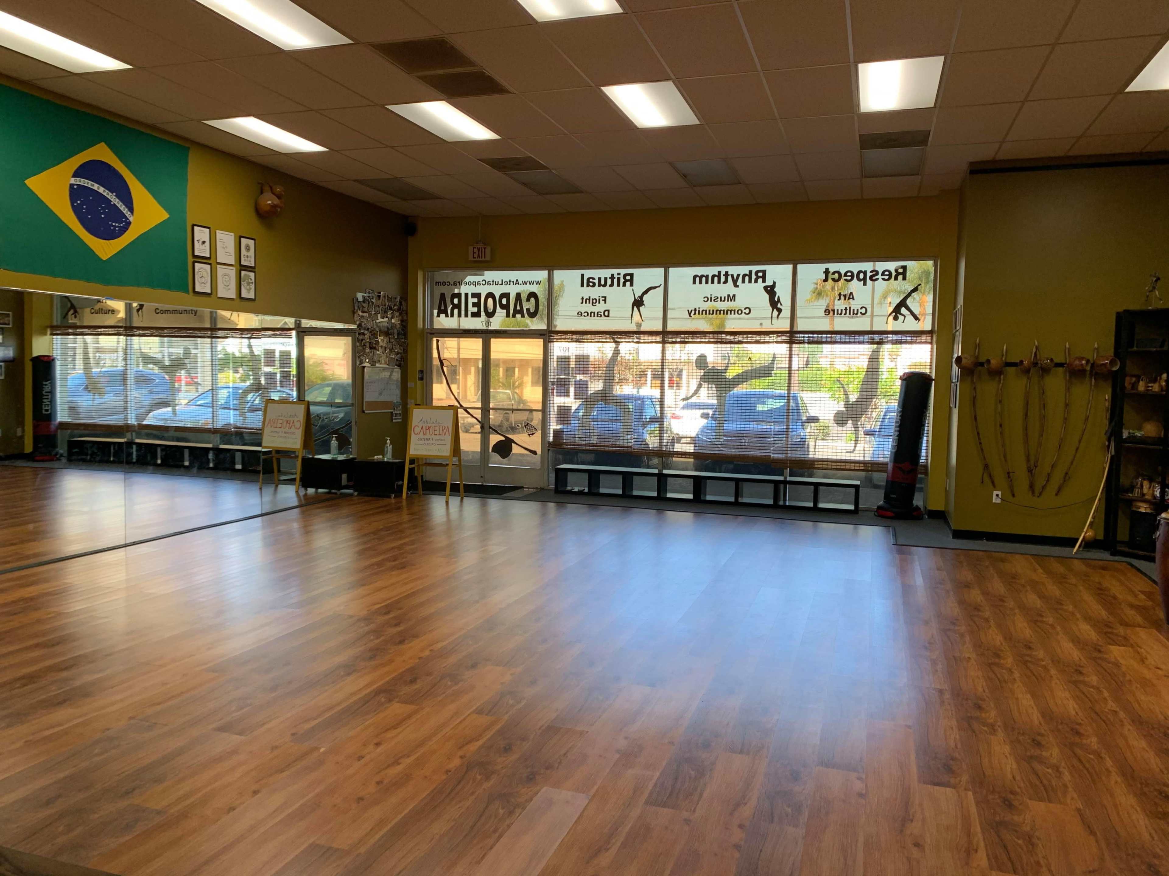 An empty fitness studio with wooden flooring and large windows displaying parked cars outside.