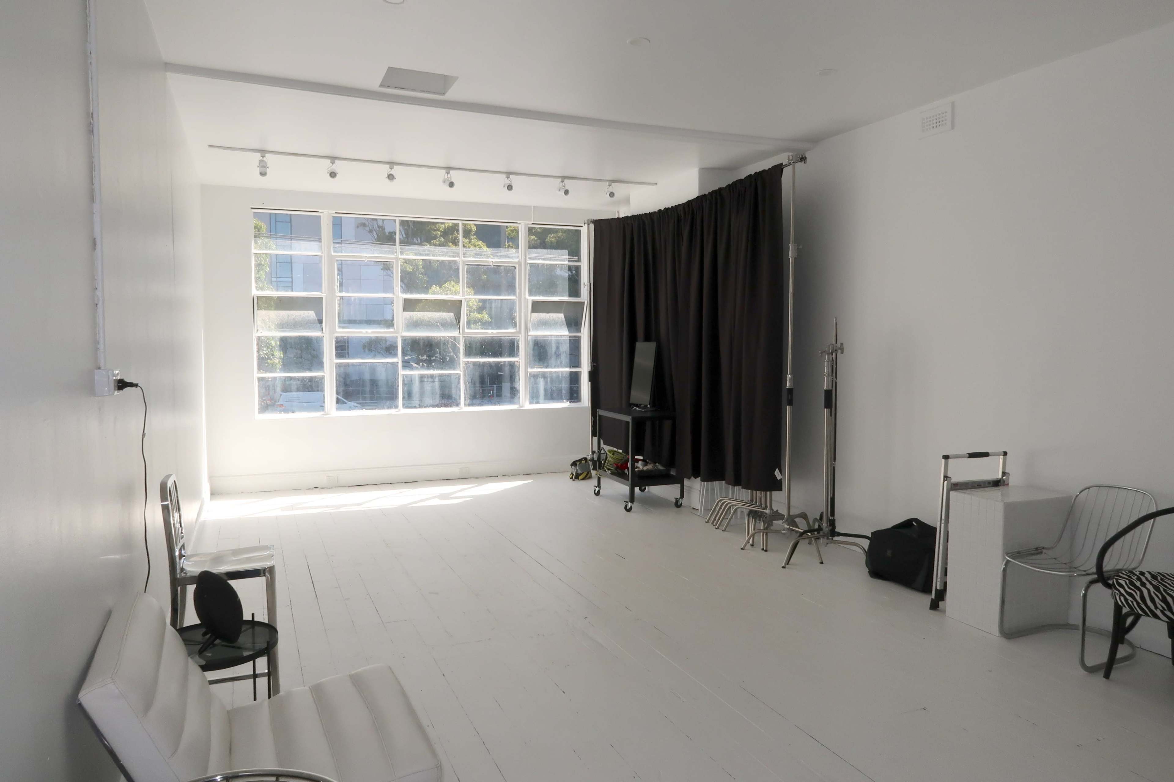 The image shows a bright, minimalist room with large windows, a sofa, a dark curtain partition, and various studio equipment.