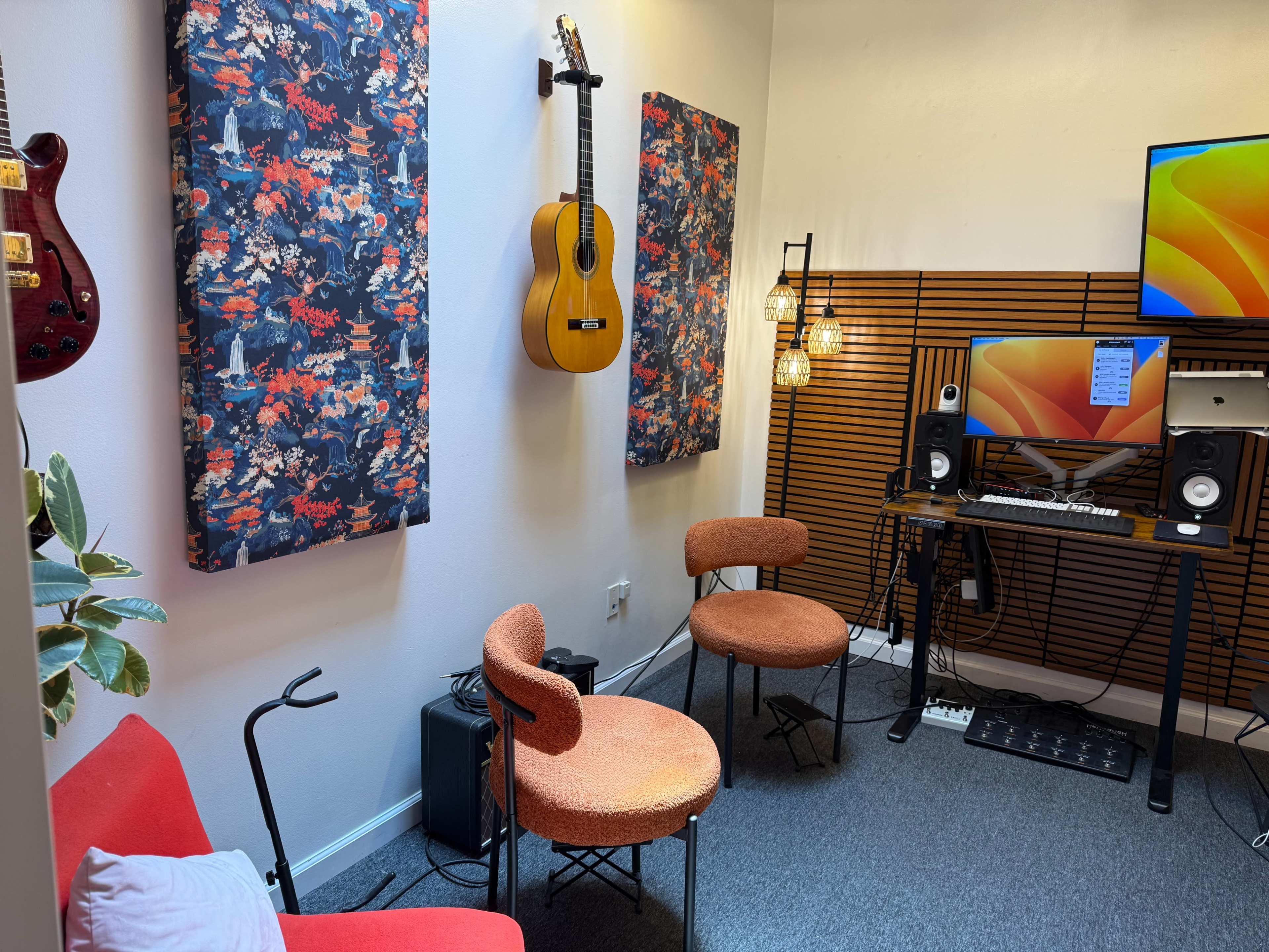 The image shows a small music studio with two colorful wall-mounted panels, a guitar, a computer setup with speakers, and three chairs arranged in a cozy arrangement.