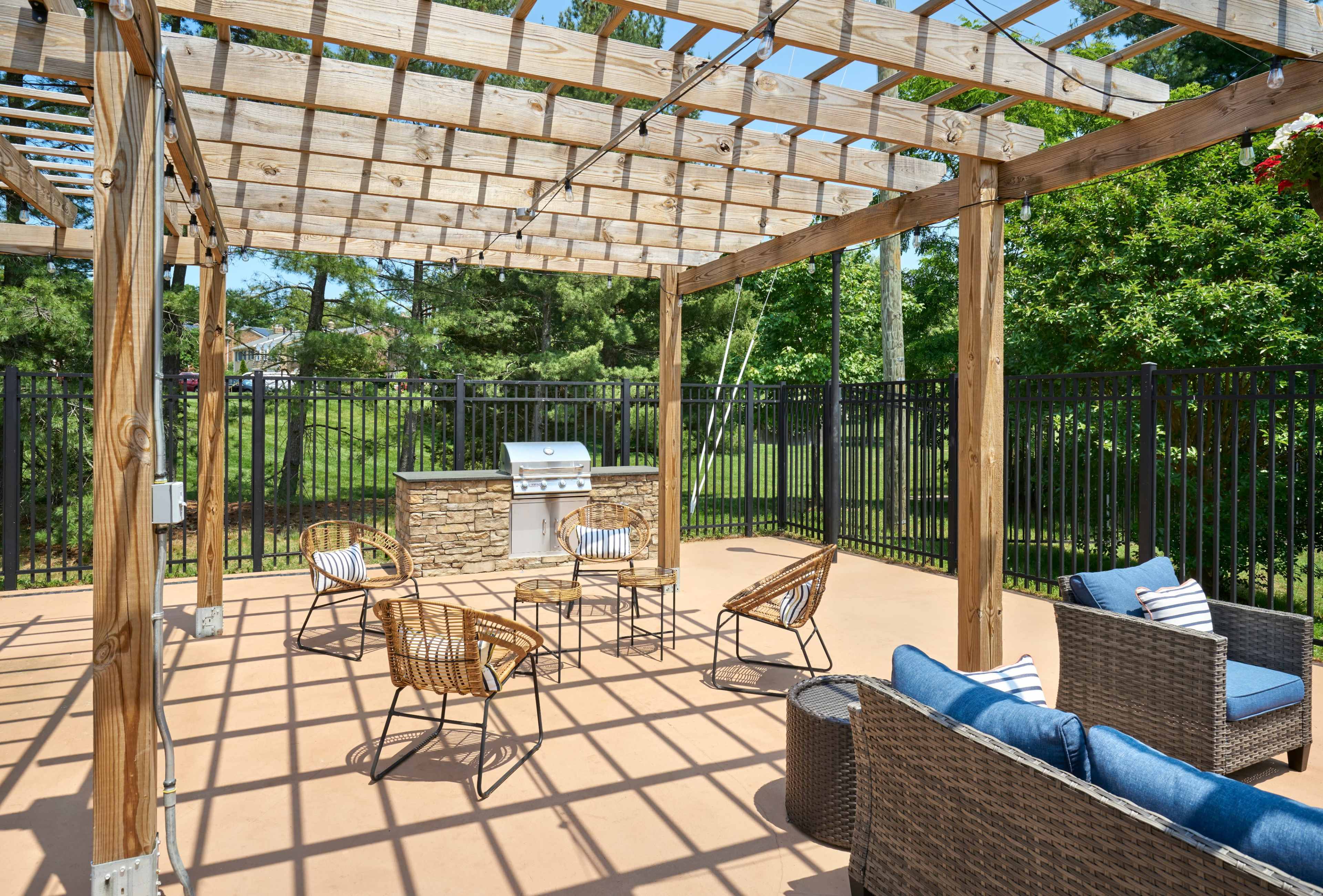 The image shows a shaded outdoor patio area with a stone barbecue grill, four wicker chairs, and a seating arrangement surrounded by a black fence and greenery.