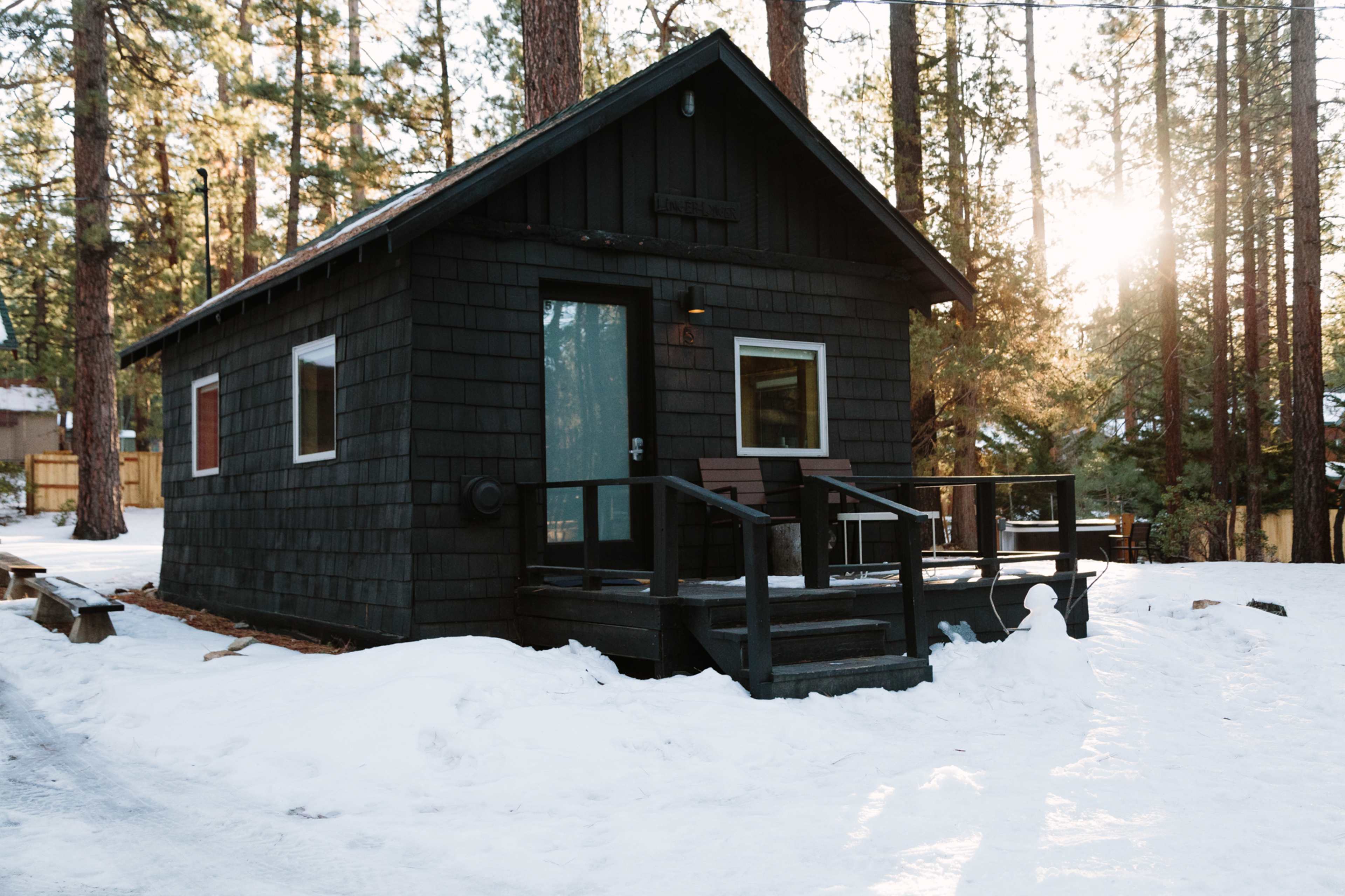 A small black cabin with a porch sits among snow-covered trees, illuminated by sunlight during winter.